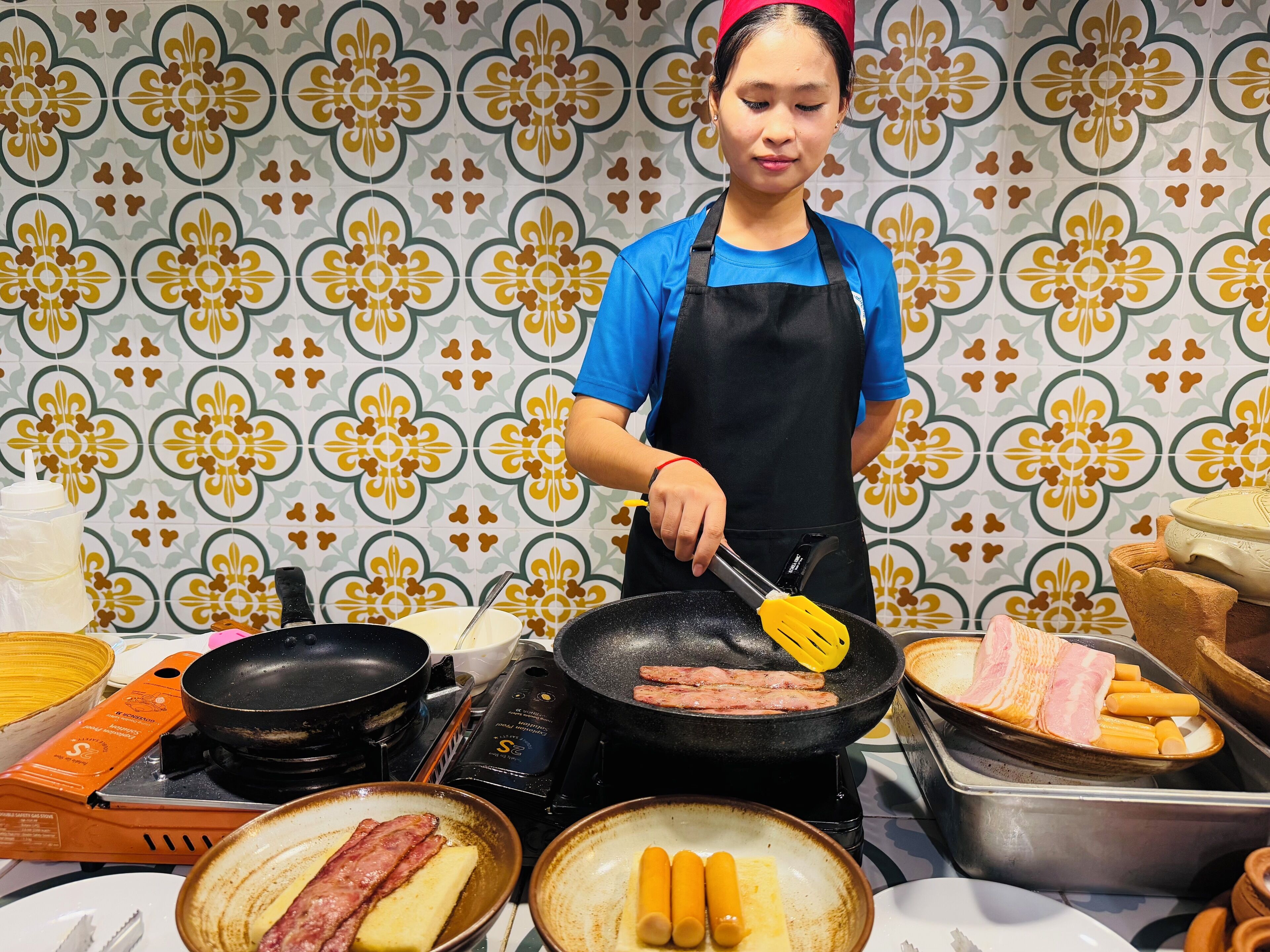 A kitchen with a person cooking sausages and bacon on a stove.