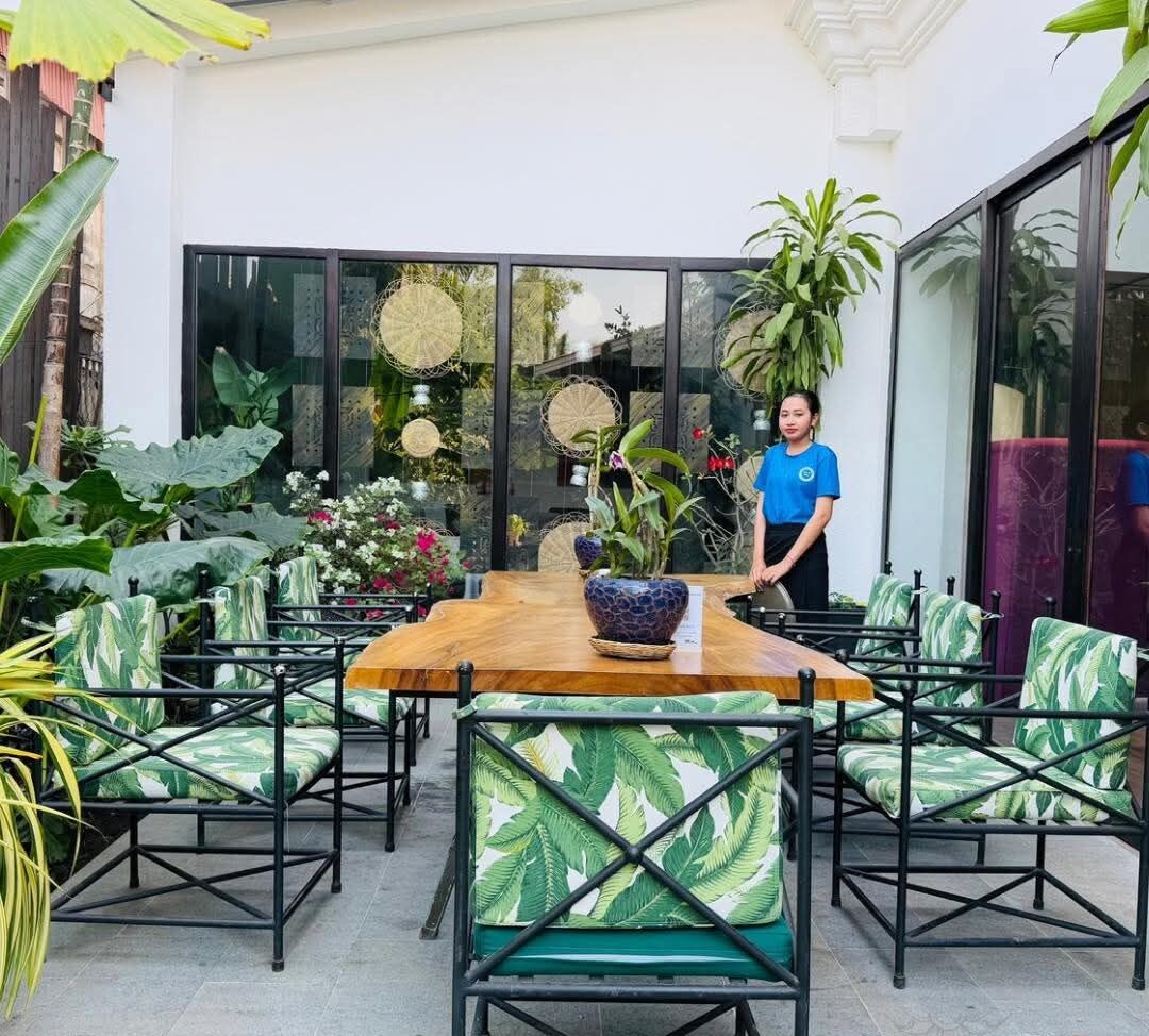 An outdoor dining area with a wooden table and chairs, surrounded by lush plants and a large window.