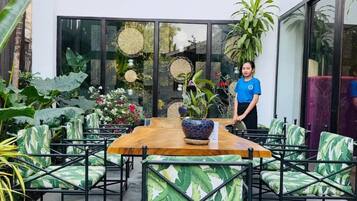 An outdoor dining area with a wooden table and chairs, surrounded by lush plants and a large window.