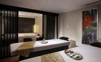 A modern hotel room with a large bed, a separate sofa, a small table with a tray of tea and snacks, a bathroom with a sink and mirror, and a large painting on the wall.