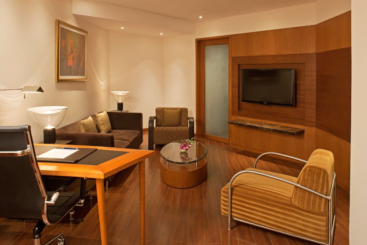 Habitación de hotel moderna con suelo de madera, una mesa de centro redonda de vidrio, un sofá, una silla, un escritorio con lámpara y un televisor de pantalla plana montado en la pared.