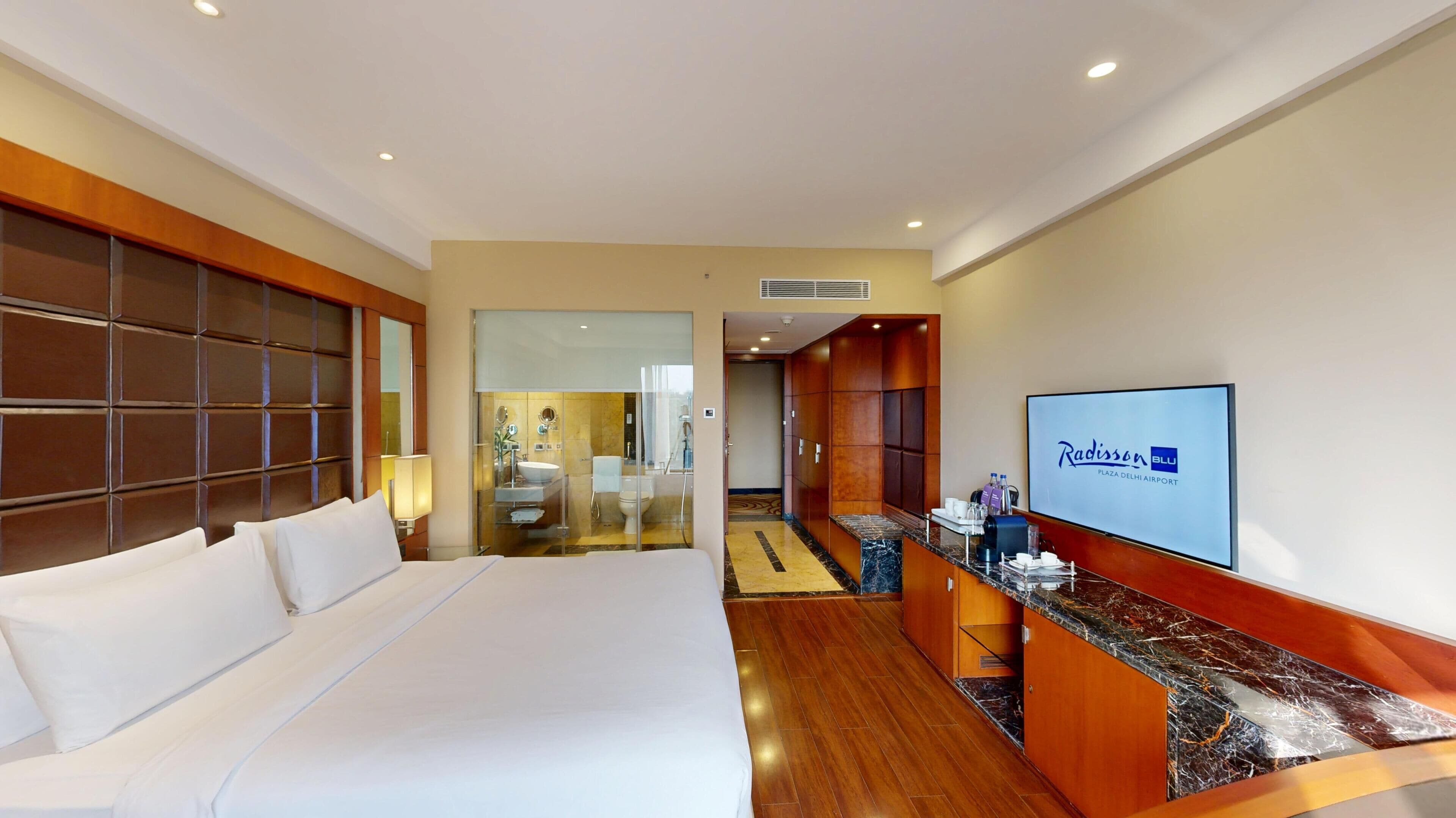 Habitación de hotel con una cama grande, televisión, minibar y baño visible a través de un separador de vidrio.
