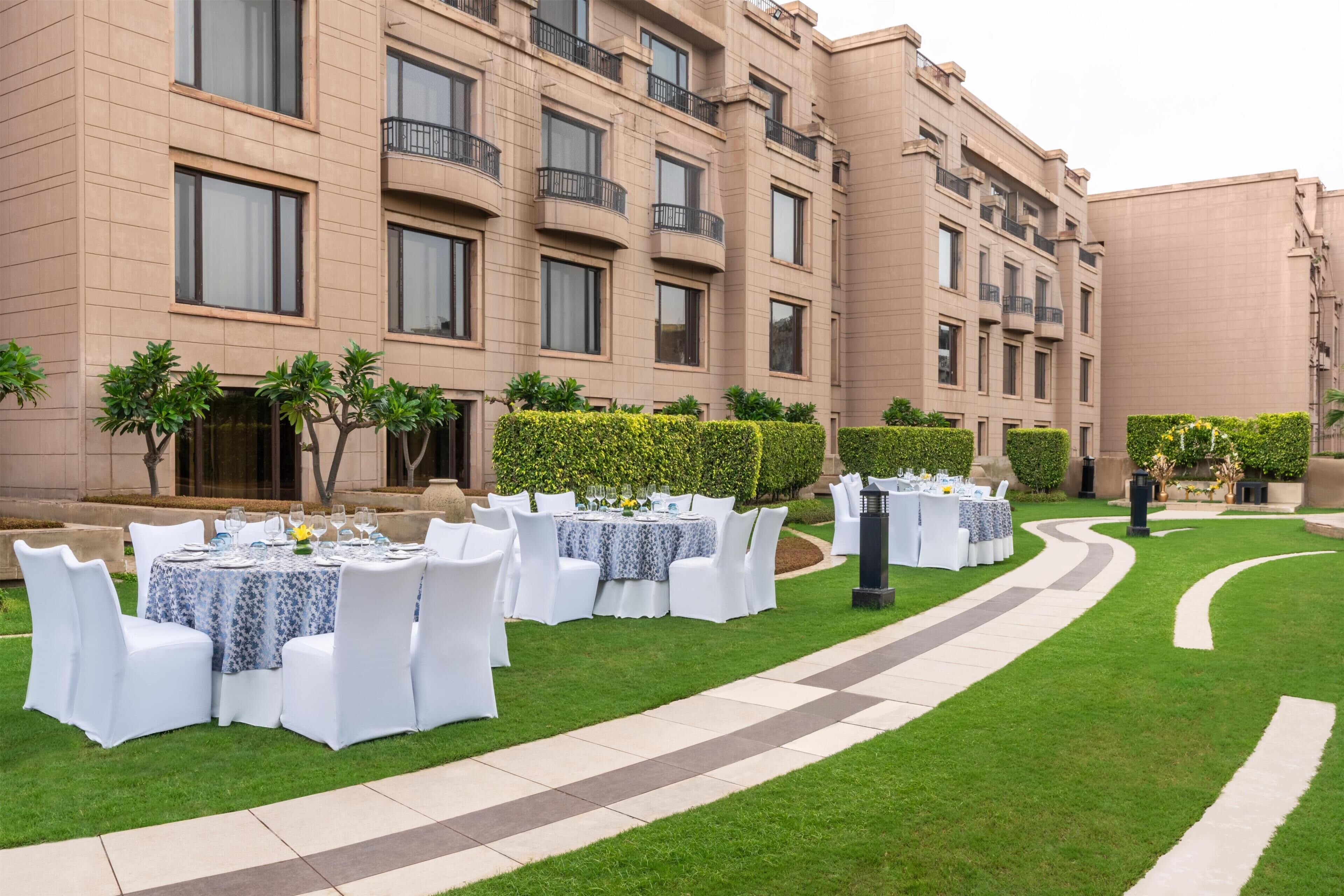 Un patio con mesas para comer al aire libre preparadas para un evento, rodeado de un césped cuidado y setos.