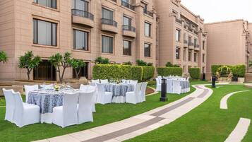 Un patio con mesas para comer al aire libre preparadas para un evento, rodeado de un césped cuidado y setos.