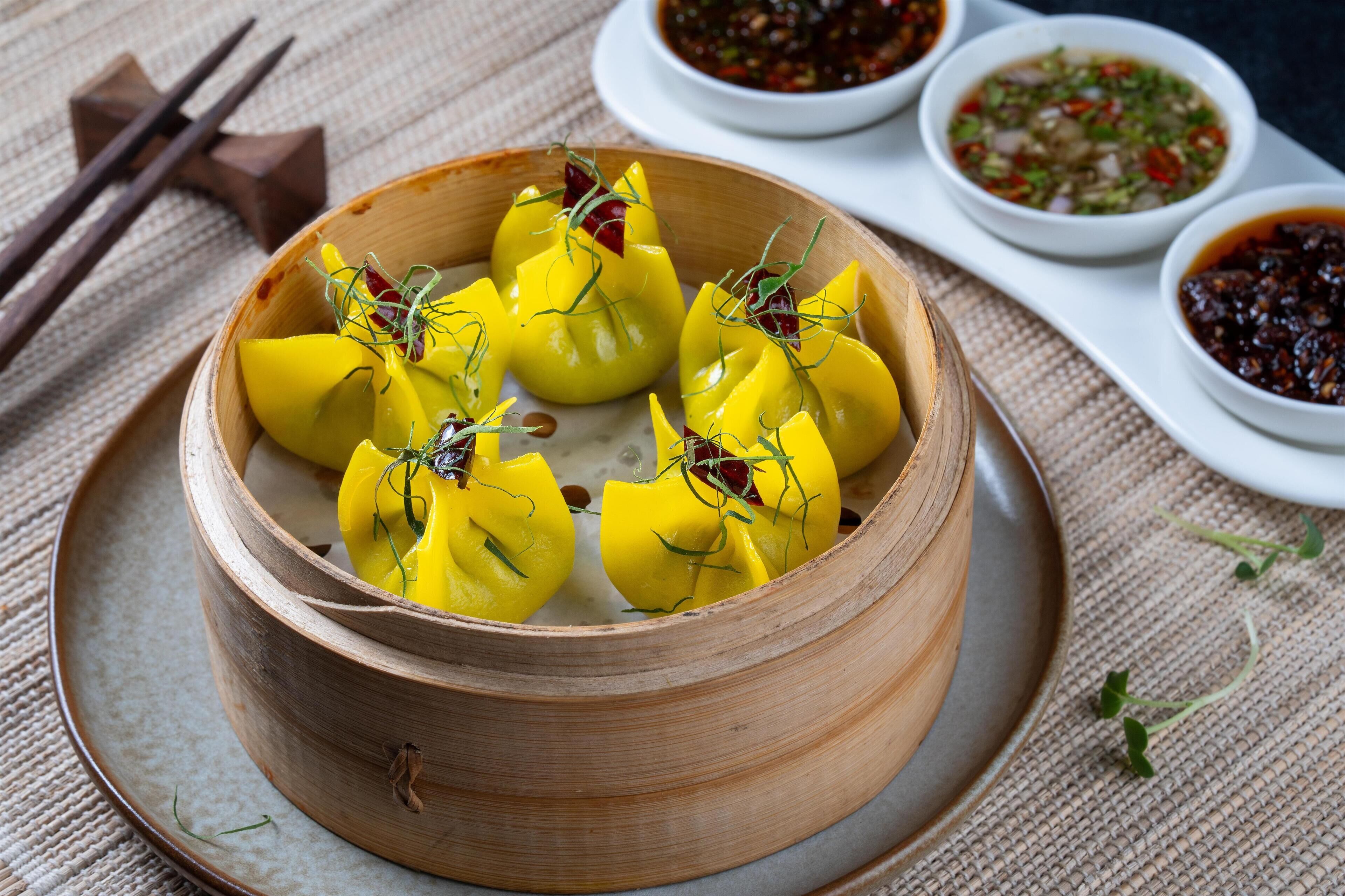 A bamboo steamer with yellow dumplings garnished with herbs, accompanied by small bowls of dipping sauces.