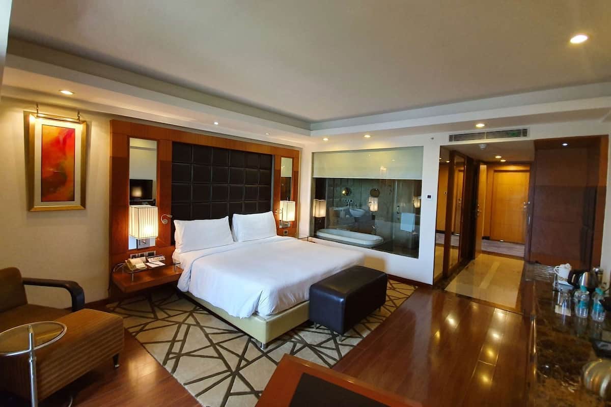 Habitación de hotel con una cama grande, mesitas de noche, un escritorio, una silla, un espejo y un baño visible a través de un panel de vidrio.