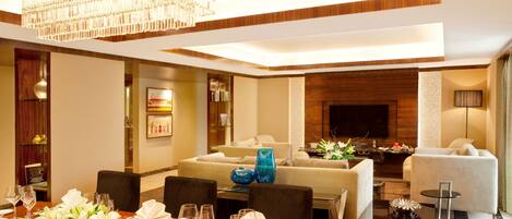 A modern hotel dining area with a wooden table set for a meal, a chandelier, and a living area with sofas and a flat-screen TV.