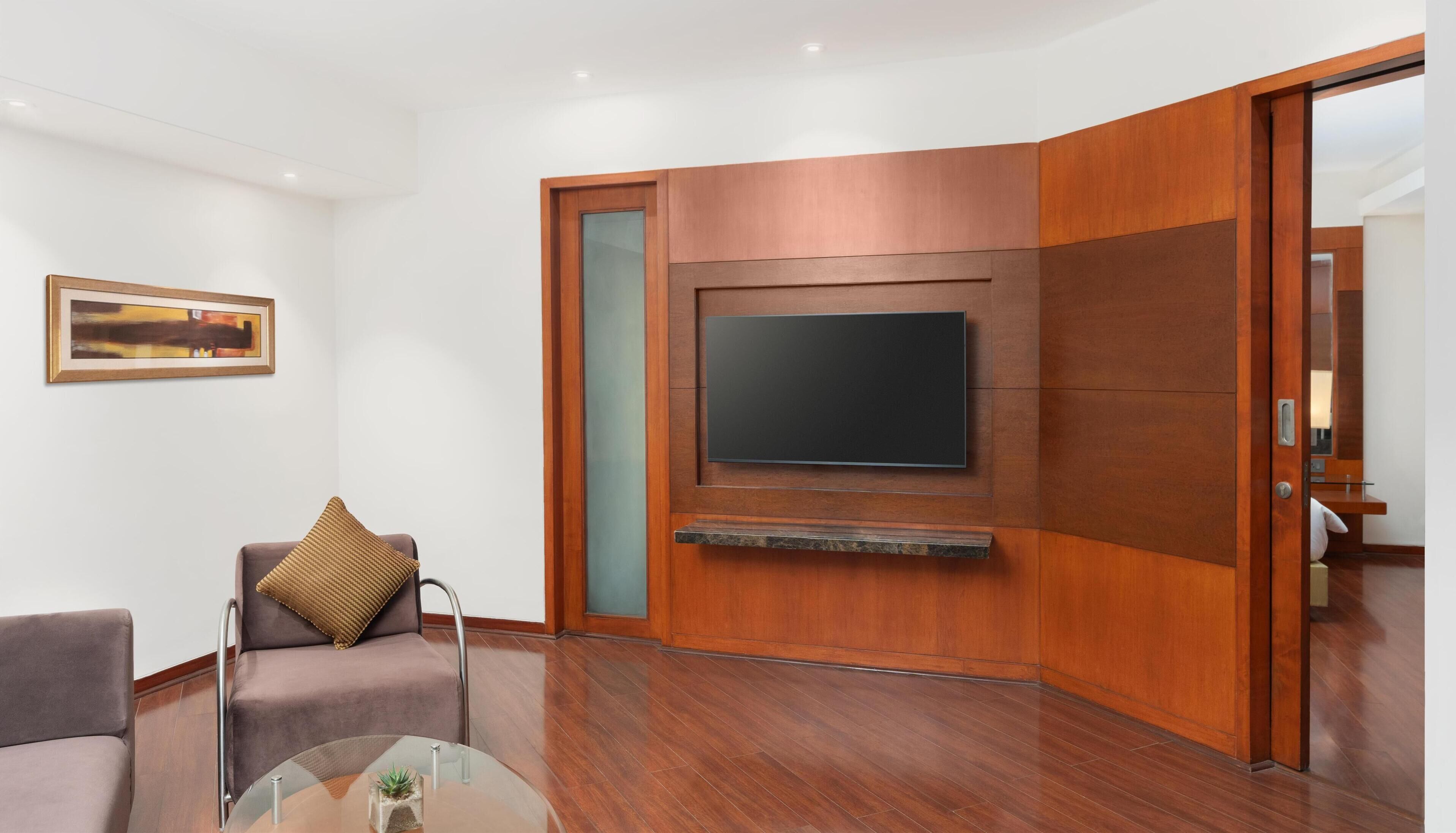 Un salon moderne avec un téléviseur à écran plat fixé sur un panneau en bois, un canapé avec un coussin, une table basse en verre et un tableau encadré accroché au mur.