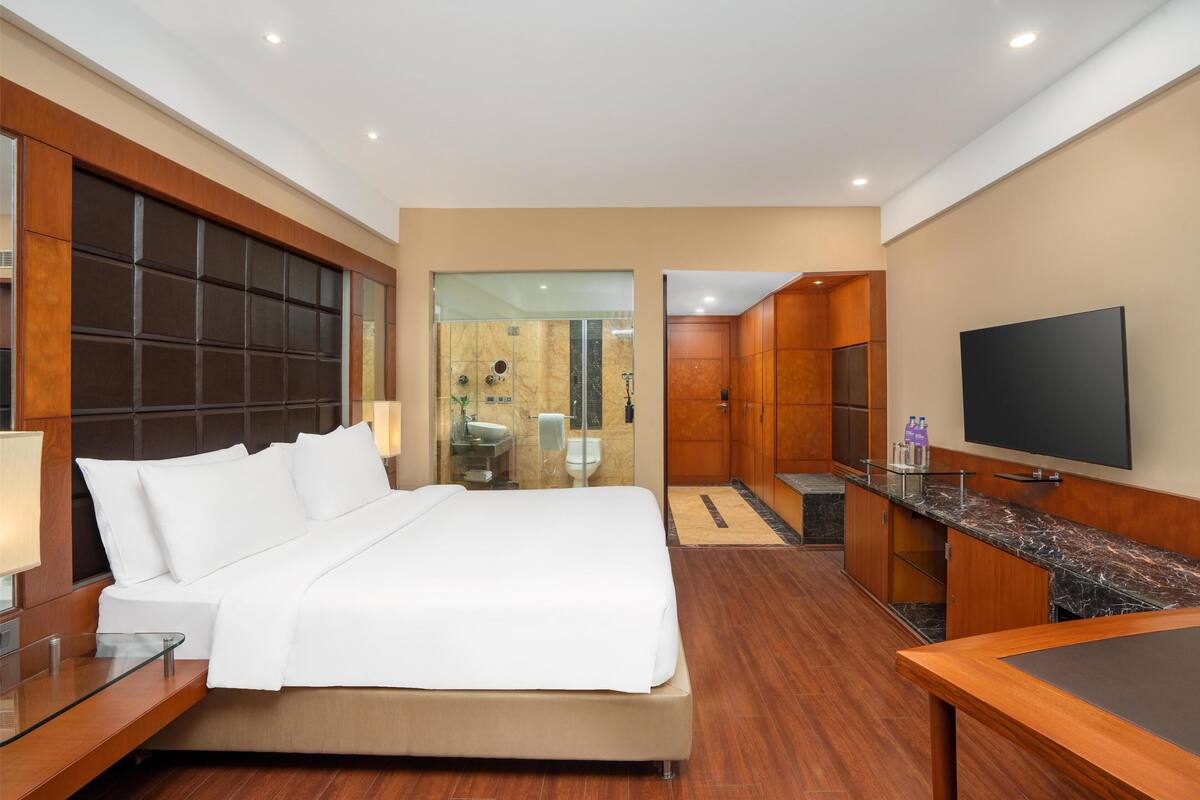Habitación de hotel con una cama grande, televisión, escritorio y baño visible a través de un panel de vidrio.