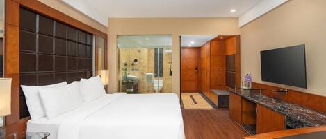 A hotel room with a large bed, a TV, a desk, and a bathroom visible through a glass partition.