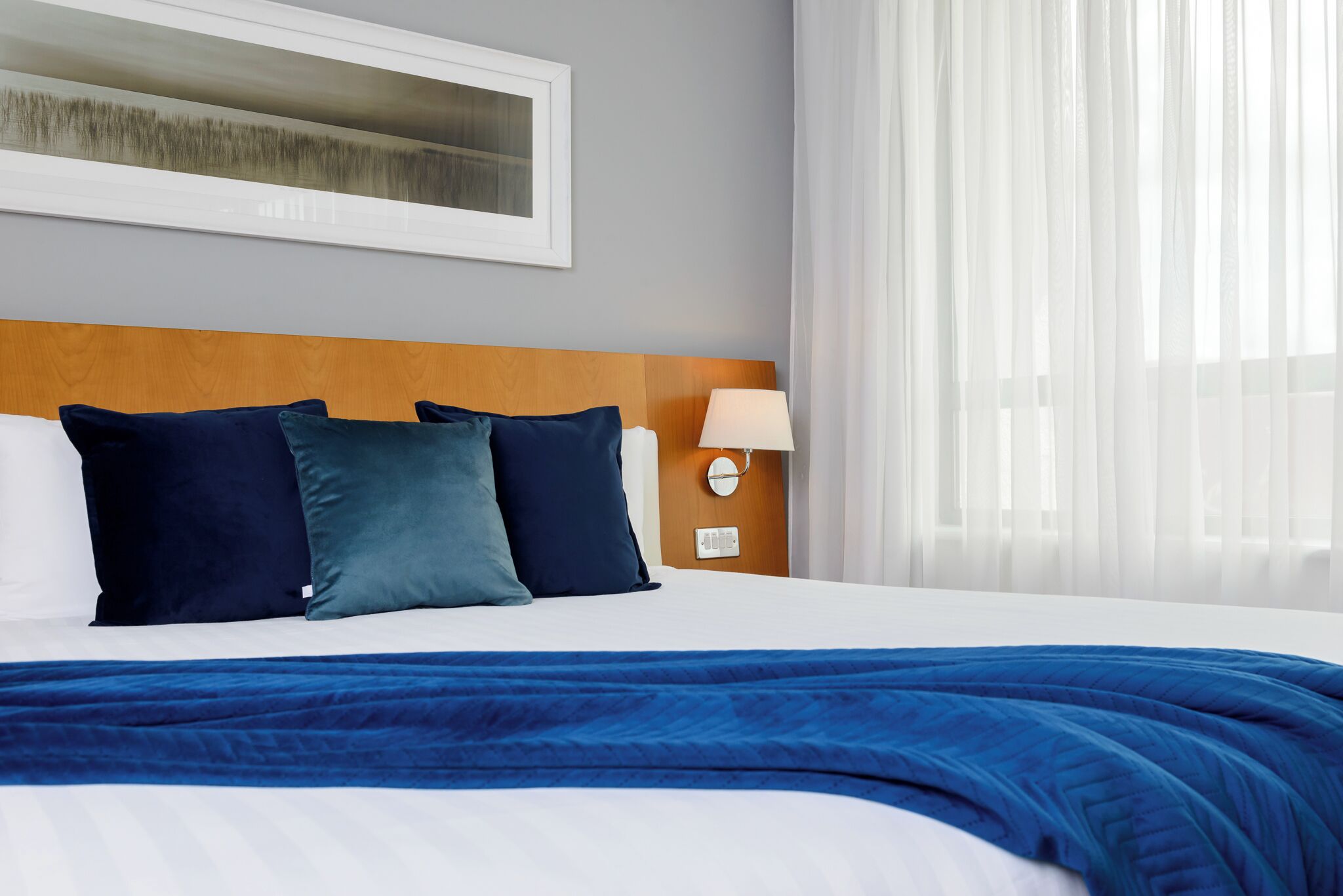 Una habitación de hotel con cama, cabecera, mesita de noche con lámpara y ventana con cortinas traslúcidas.