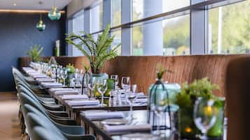 A restaurant with long tables set for dining, large windows, and modern pendant lighting.