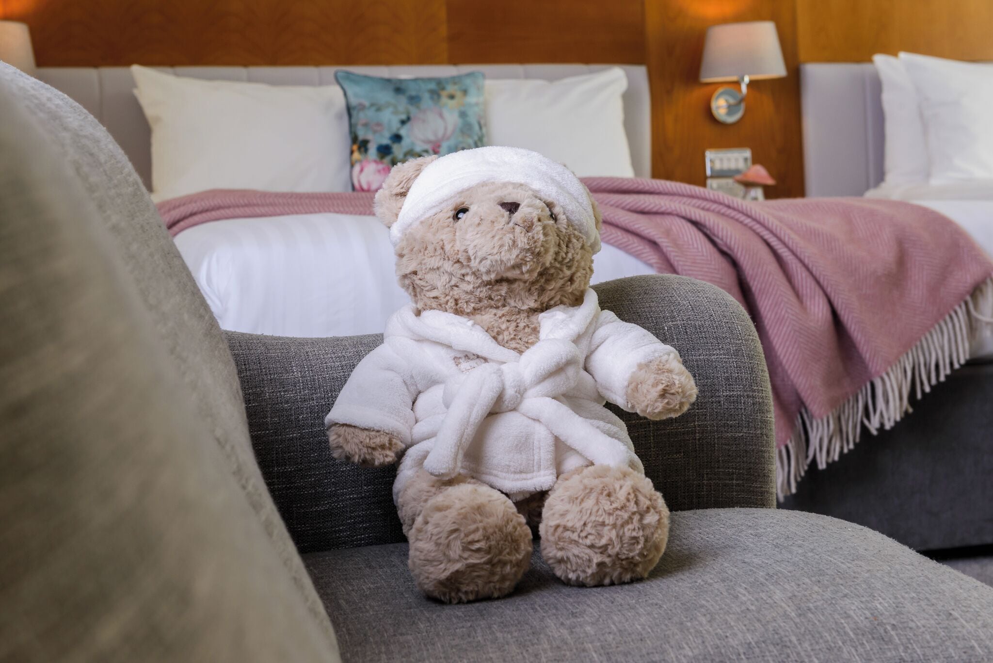 A plush teddy bear in a bathrobe sitting on a grey couch in a hotel room.