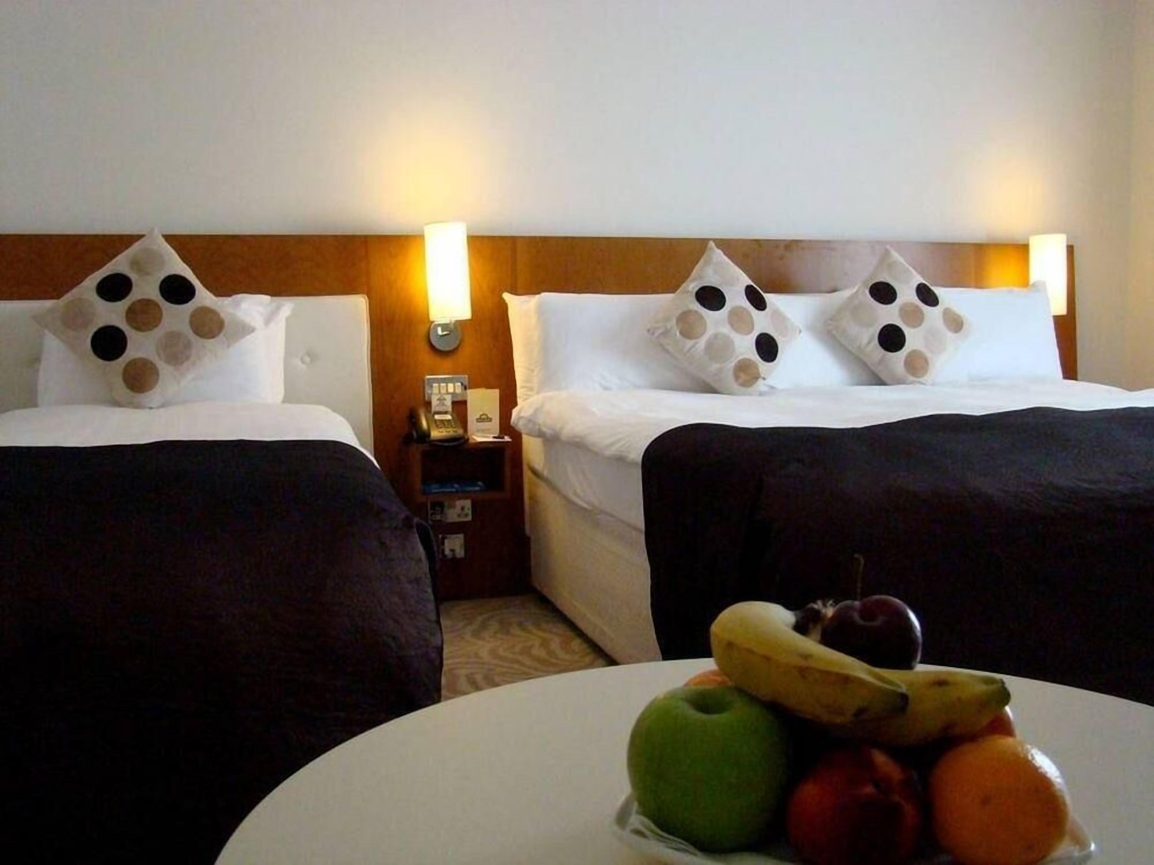 Two beds with unique pillow designs, a bedside table with a lamp, and a fruit bowl on a table.