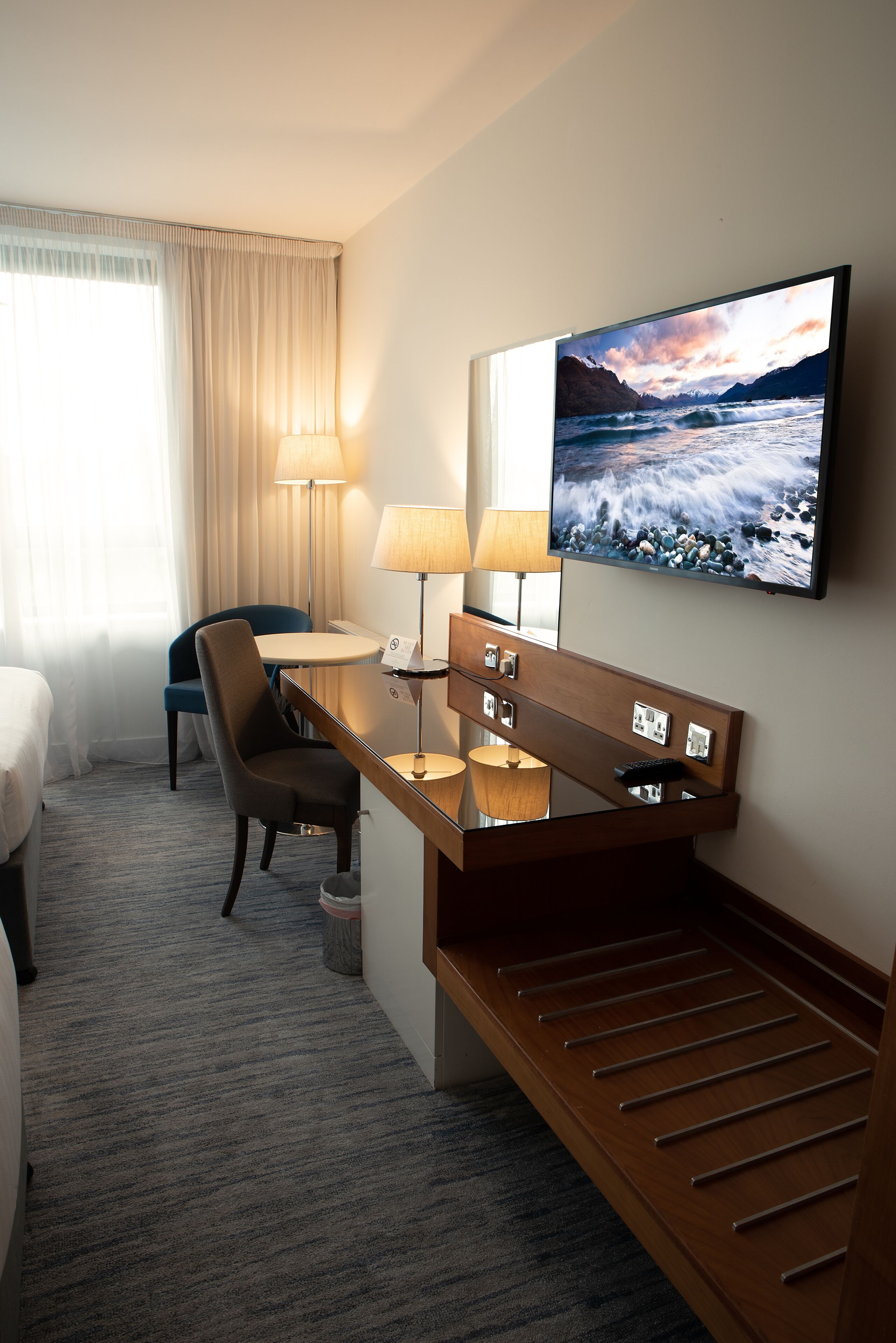 A hotel room with a bed, a desk with a lamp, a chair, a TV on the wall, and a window with curtains.