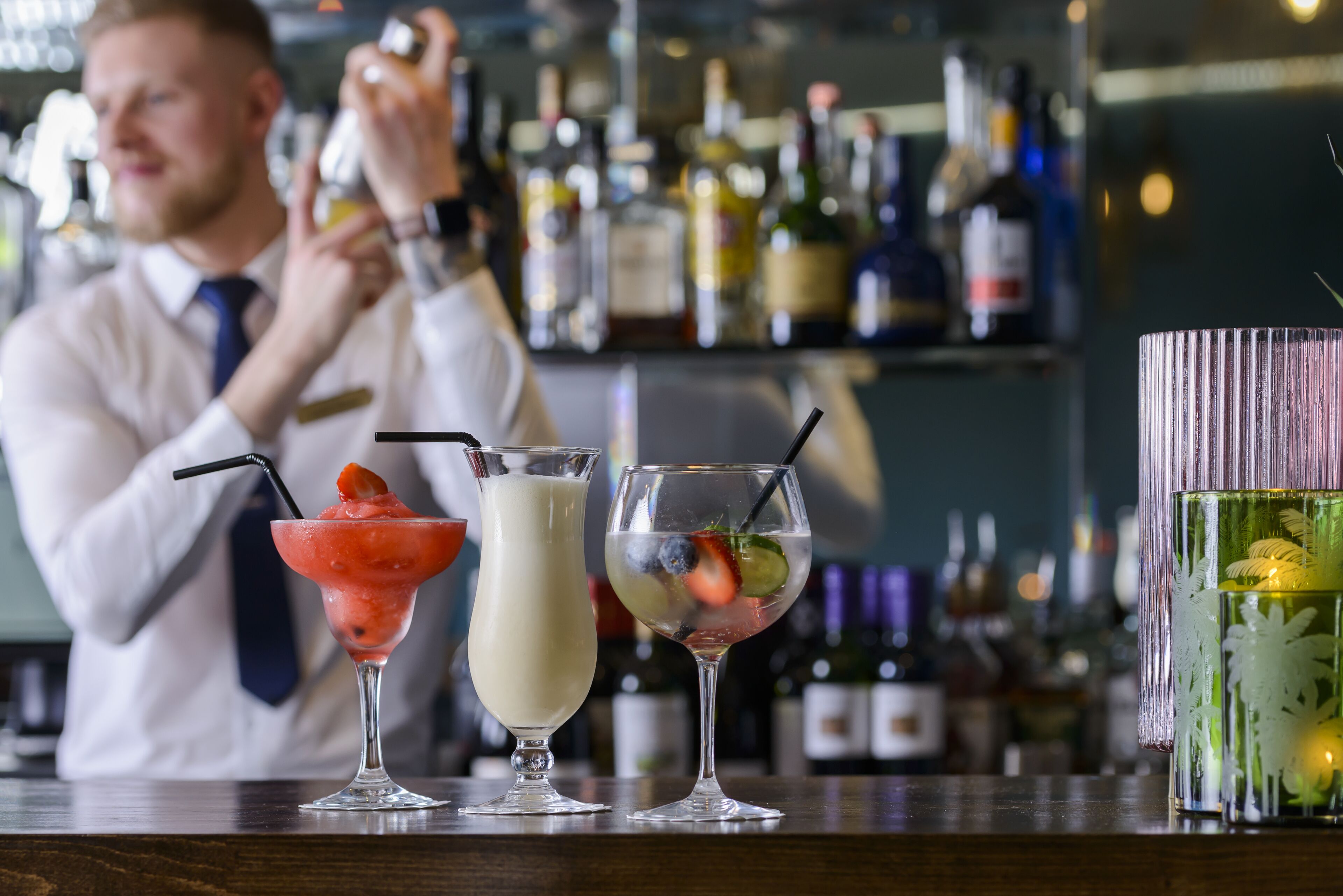 Un barman preparando bebidas detrás de la barra, con varias botellas y un recipiente de vidrio decorativo.