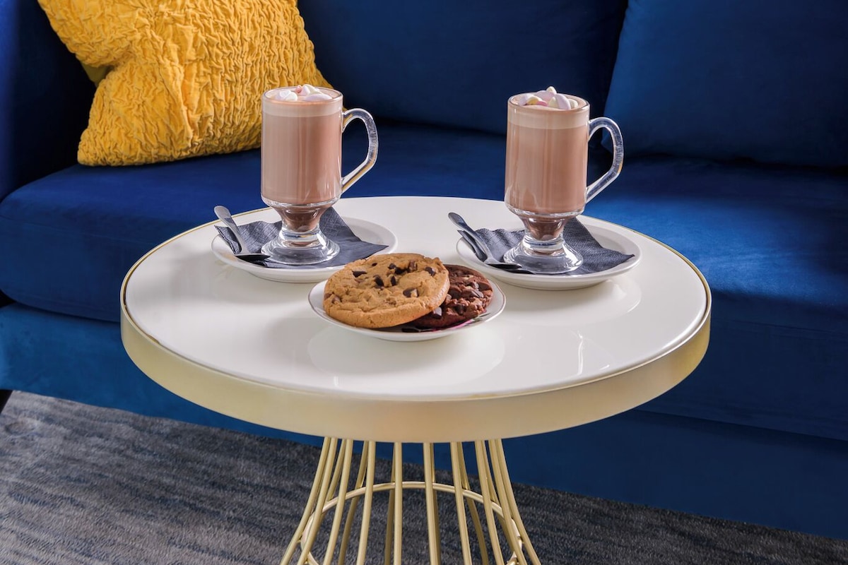 A blue sofa with yellow pillows, a white table with two mugs of hot chocolate, a plate of cookies, and a vase with white flowers on a grey wall.