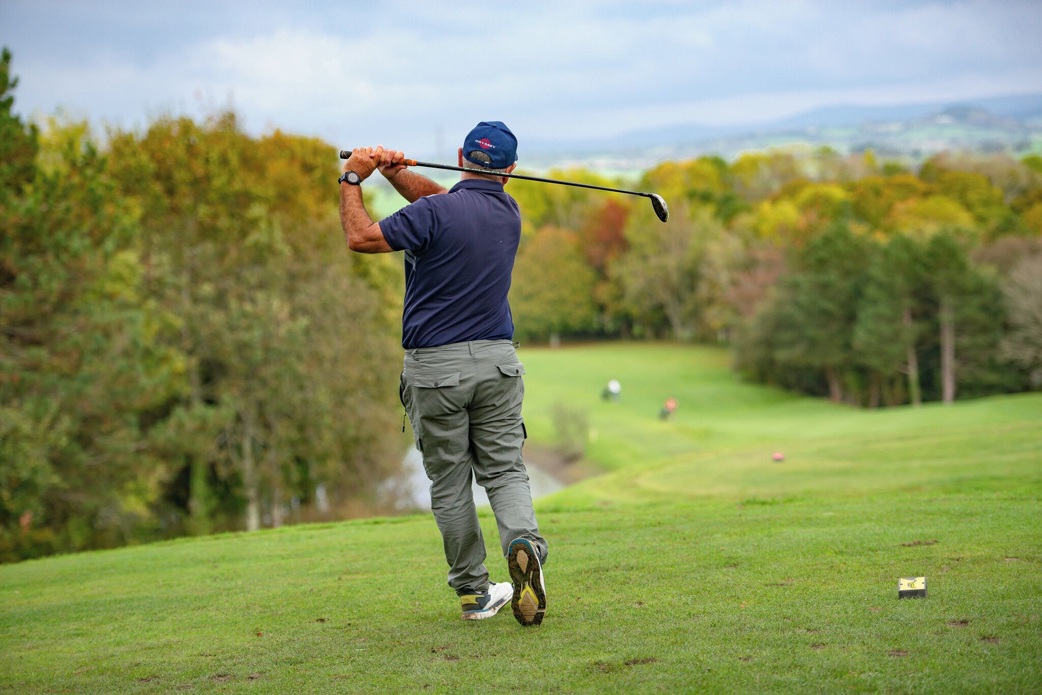 Un hombre con camisa y gorra azul balancea un palo de golf en un campo, con un río y árboles al fondo.