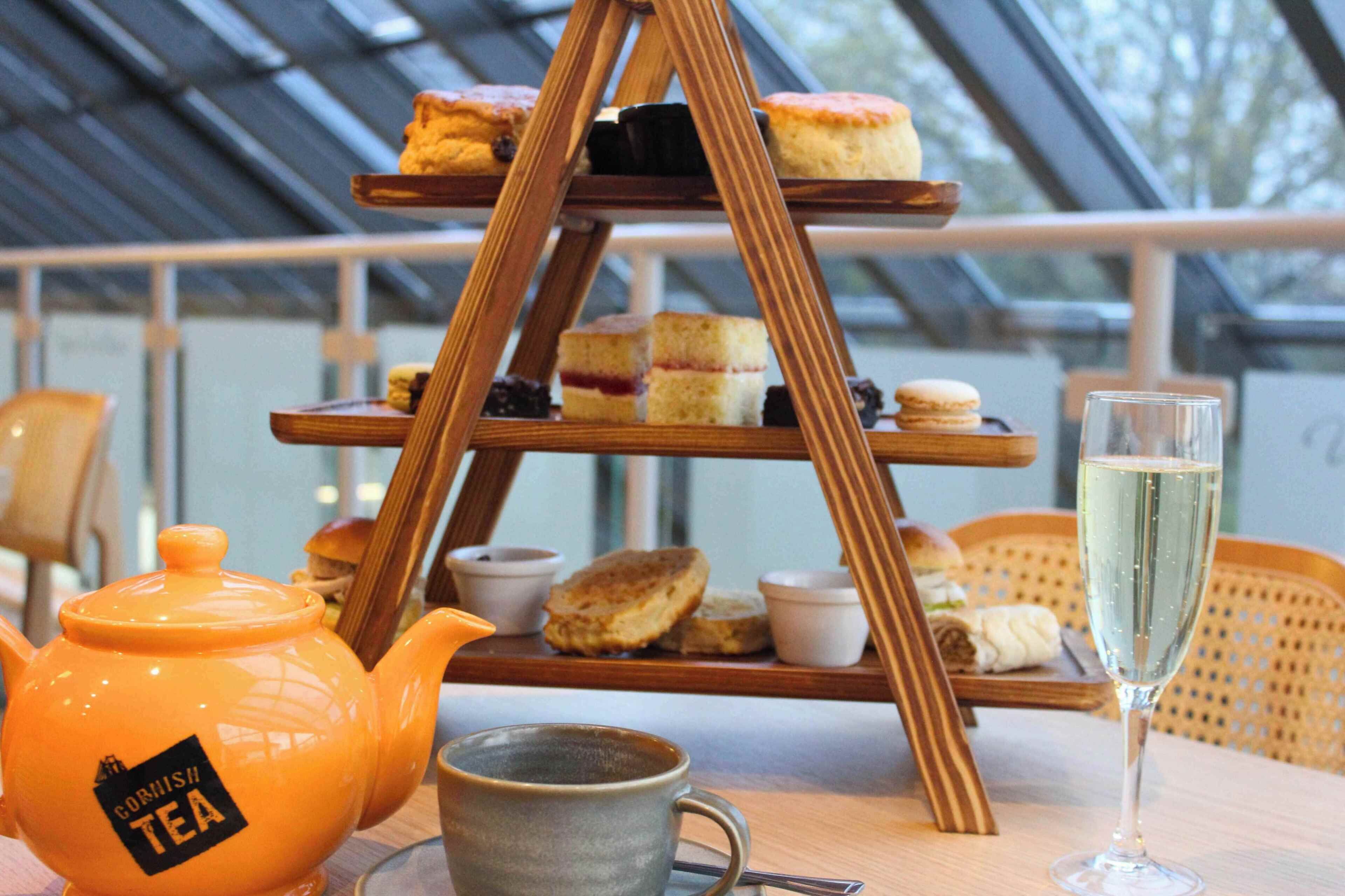 Exibição escalonada de sobremesas com diversos doces e um bule com a inscrição 'Cornish Tea' sobre uma mesa.