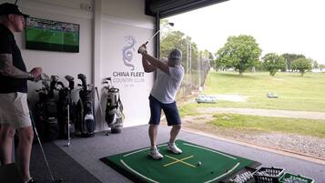Área de treino de golfe com putting green, sacos de golfe e um muro com o texto 'CHINA FLEET COUNTRY CLUB EST. 1991'.