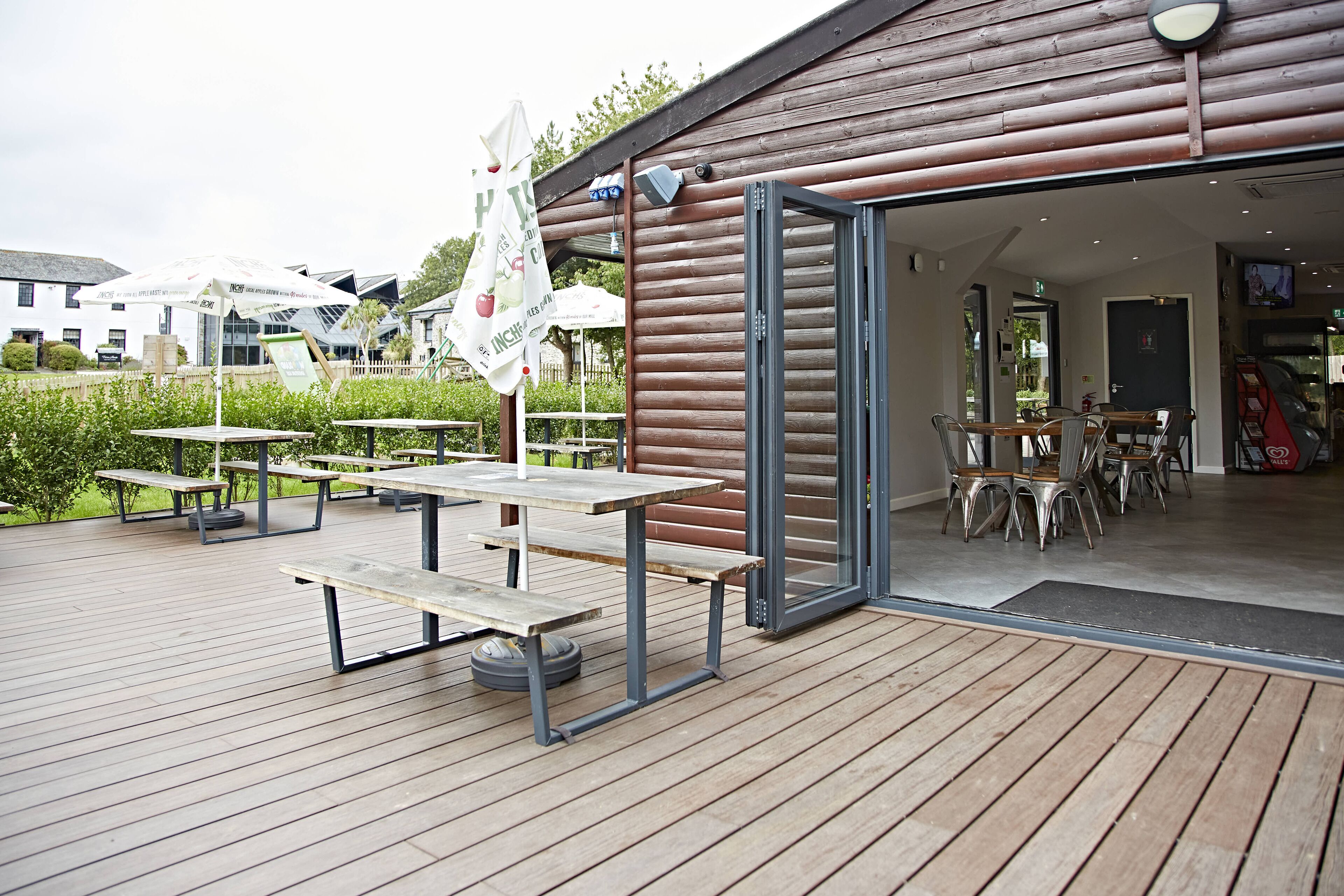 Une terrasse extérieure en bois, dotée de tables de pique-nique et d’une porte vitrée donnant accès à une salle à manger intérieure.