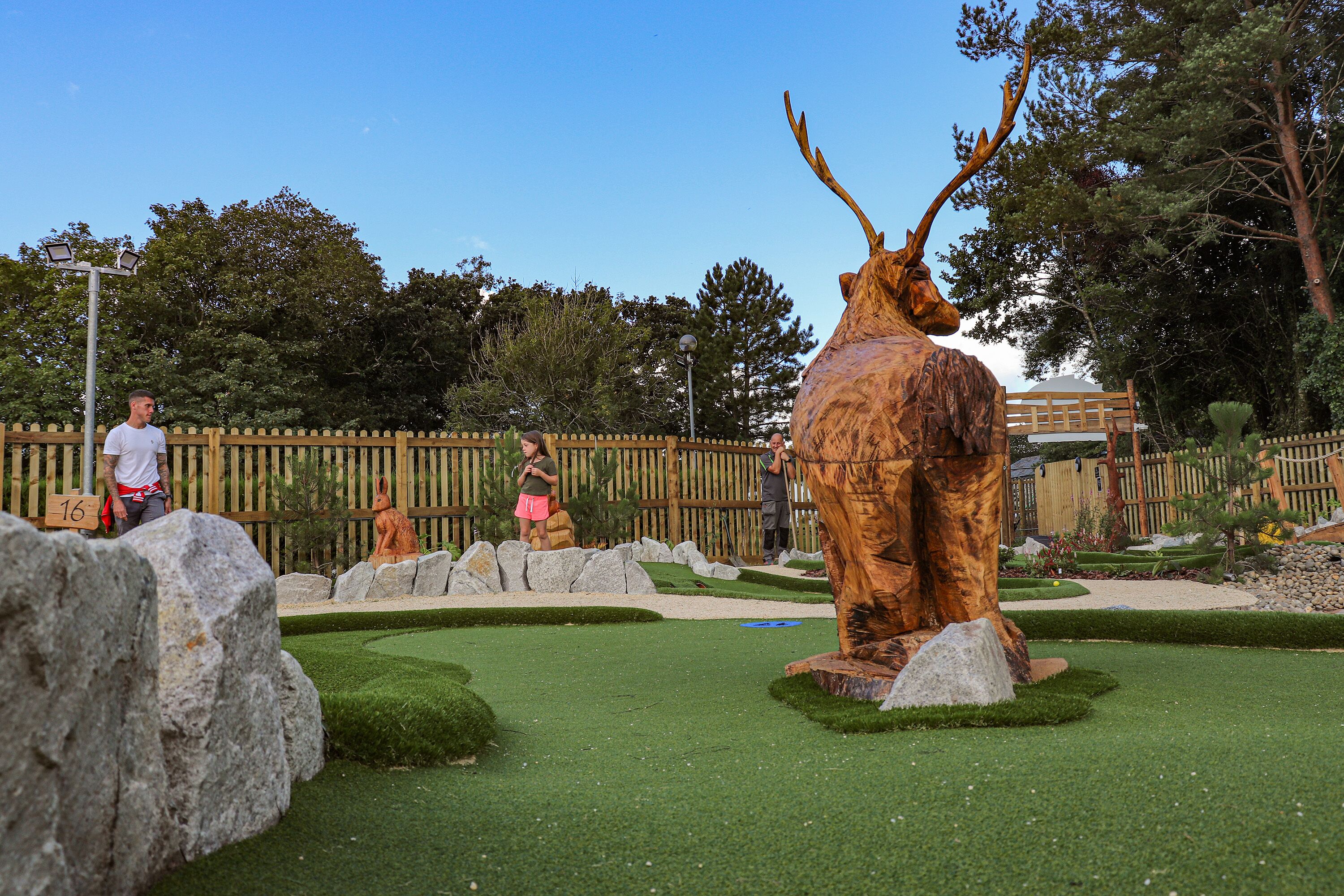 A golf course with a large deer statue, a wooden fence, and a clear blue sky.