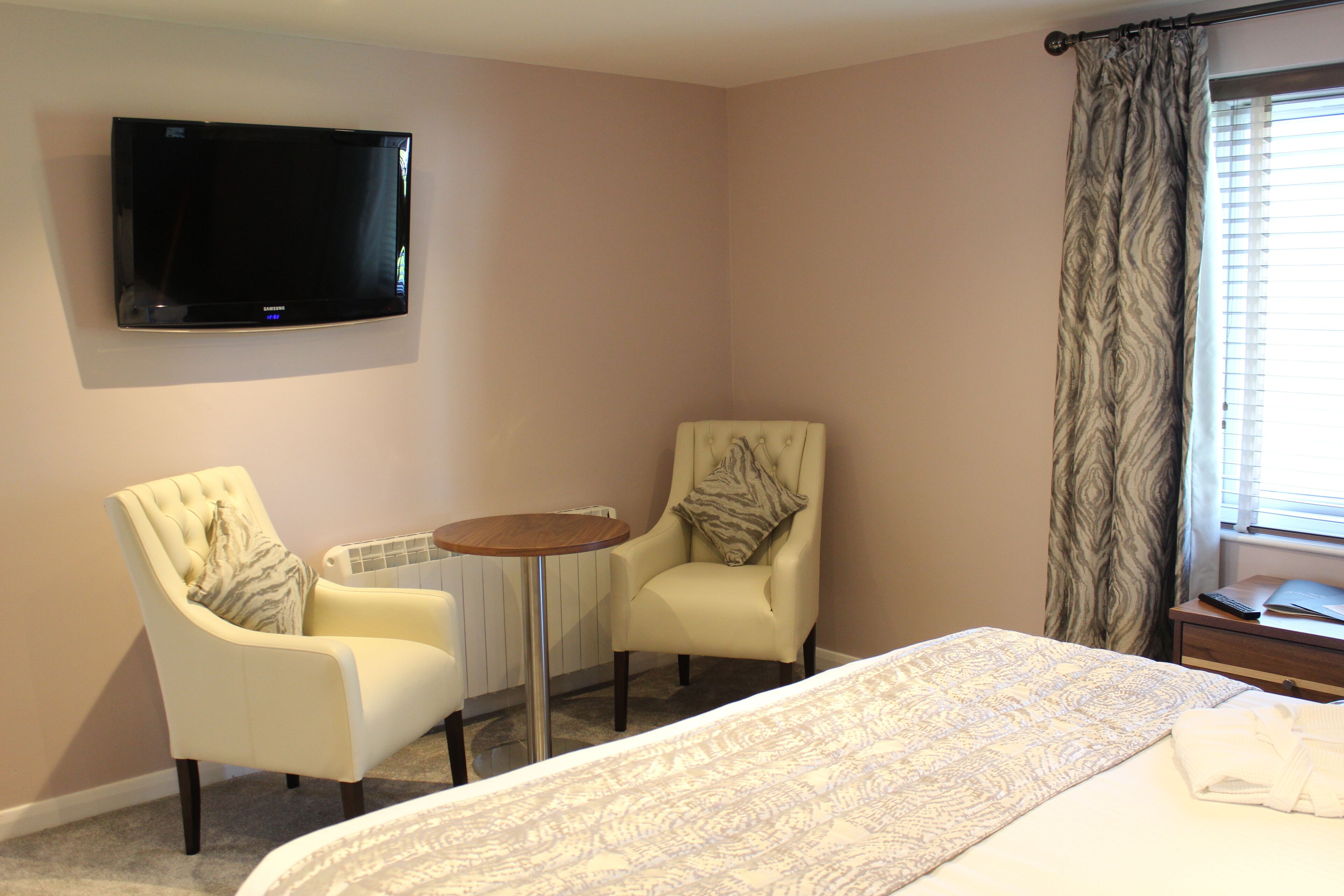 A hotel room with a bed, two armchairs, a small table, a flat-screen TV mounted on the wall, and a window with curtains.