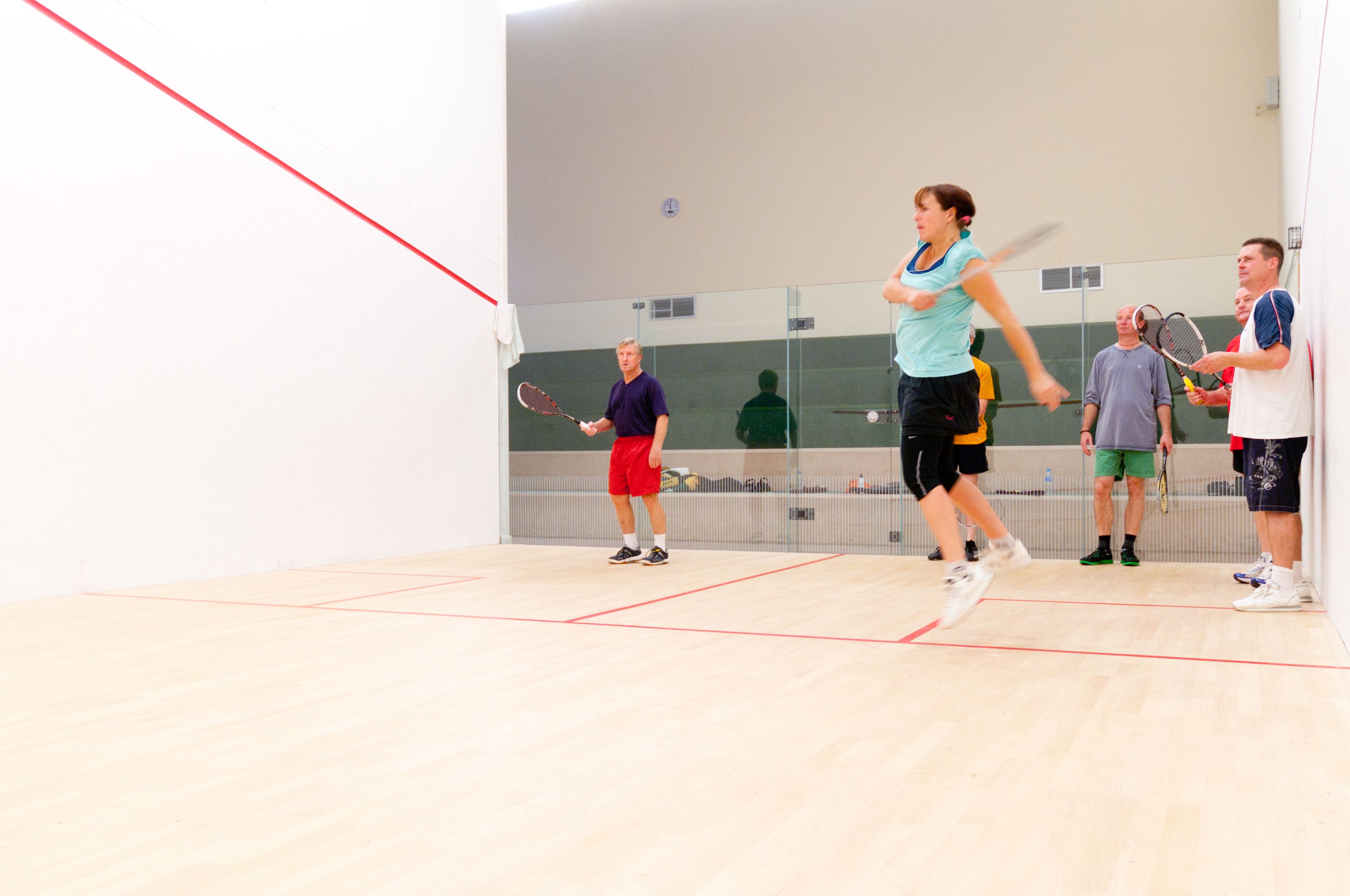 Una cancha de squash con jugadores, un reloj en la pared y un espejo que refleja la cancha.