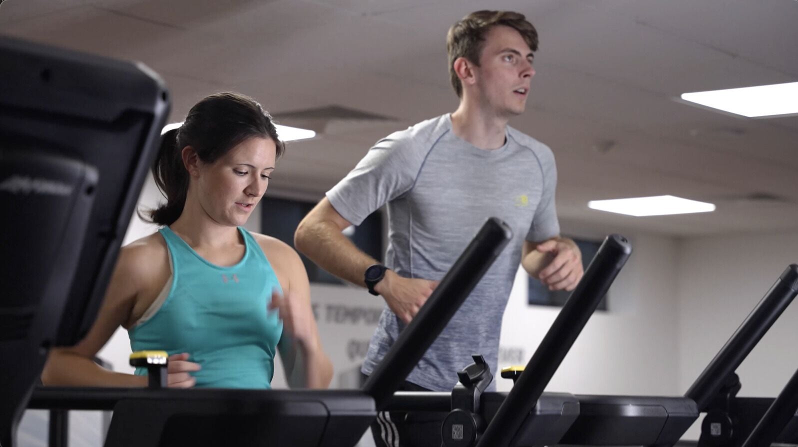 Dos personas corriendo en cintas de correr dentro de un gimnasio con equipo visible y un techo con luces.