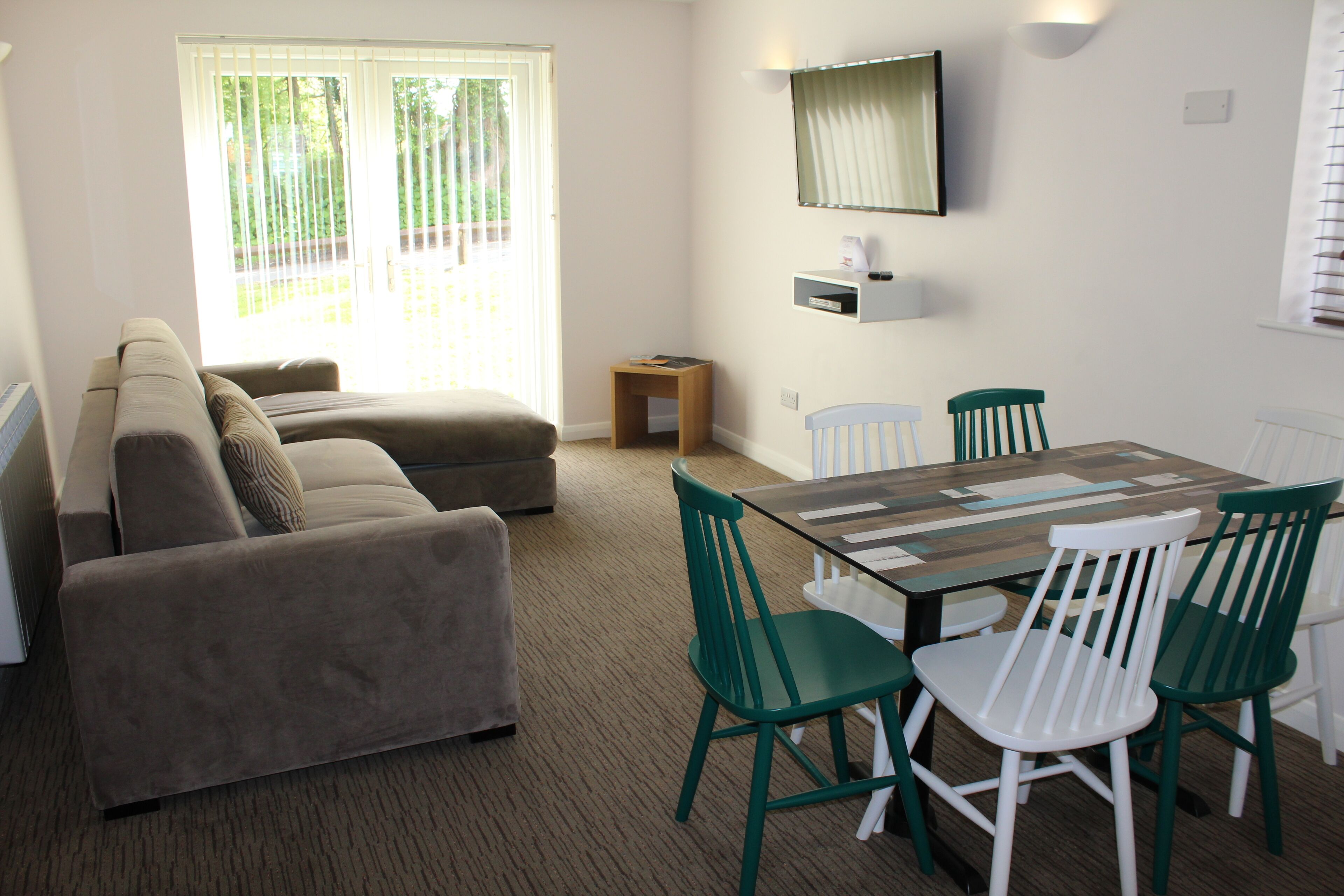 A living room with a sofa, a dining table with chairs, a small wooden stool, and a wall-mounted shelf.