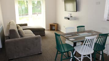 Ein Wohnzimmer mit einer Couch, einem Esstisch mit Stühlen, einem kleinen Holzschemel und einem an der Wand befestigten Regal.
