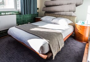 Una cama bien hecha con una manta gris, cabecera de madera y una mesita de noche con una lámpara y una caja de pañuelos.