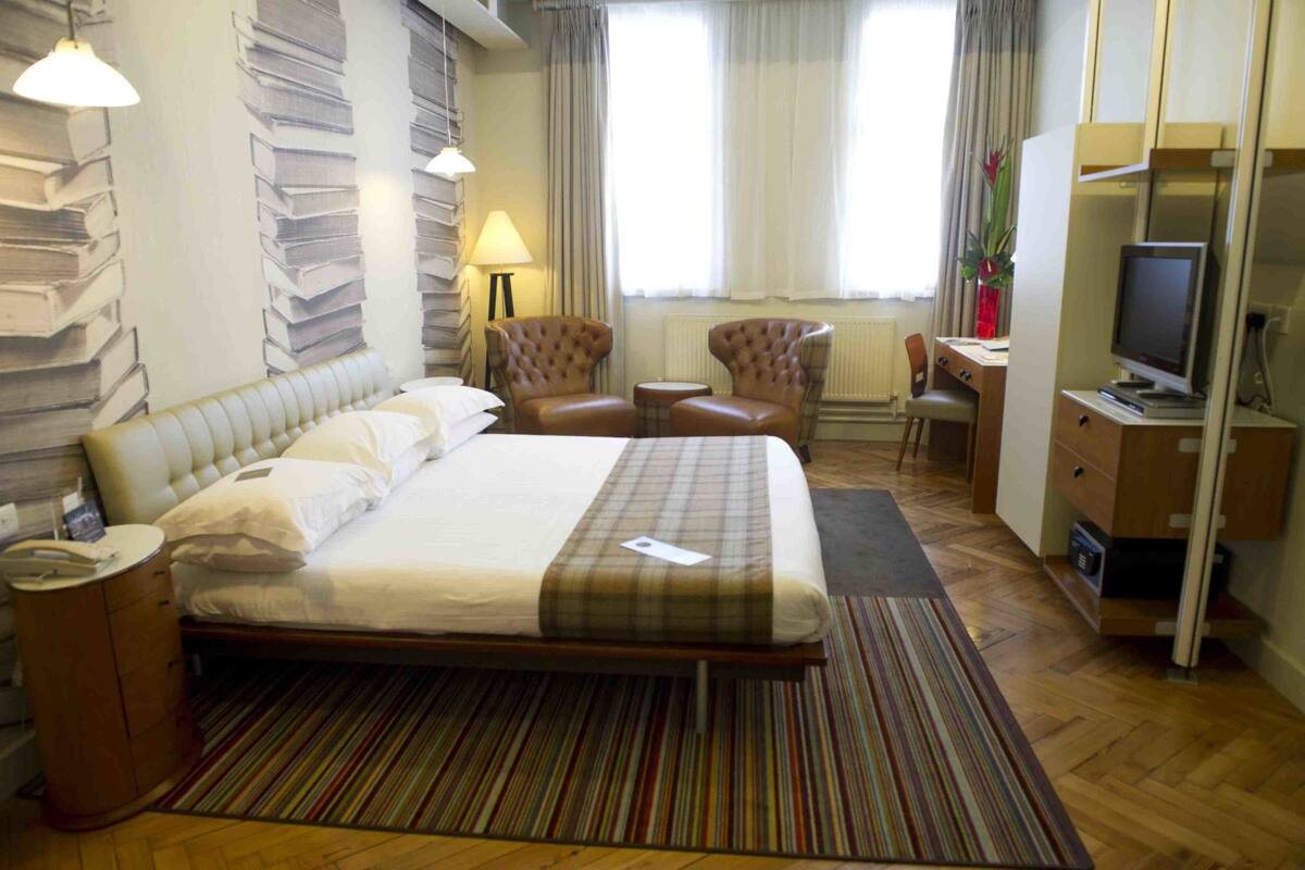 A hotel room with a large bed, a desk with a computer, two armchairs, a small table, and a TV.