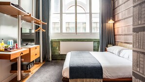 Una camera d'albergo con un letto, una scrivania con la televisione, una libreria e una finestra con tende.