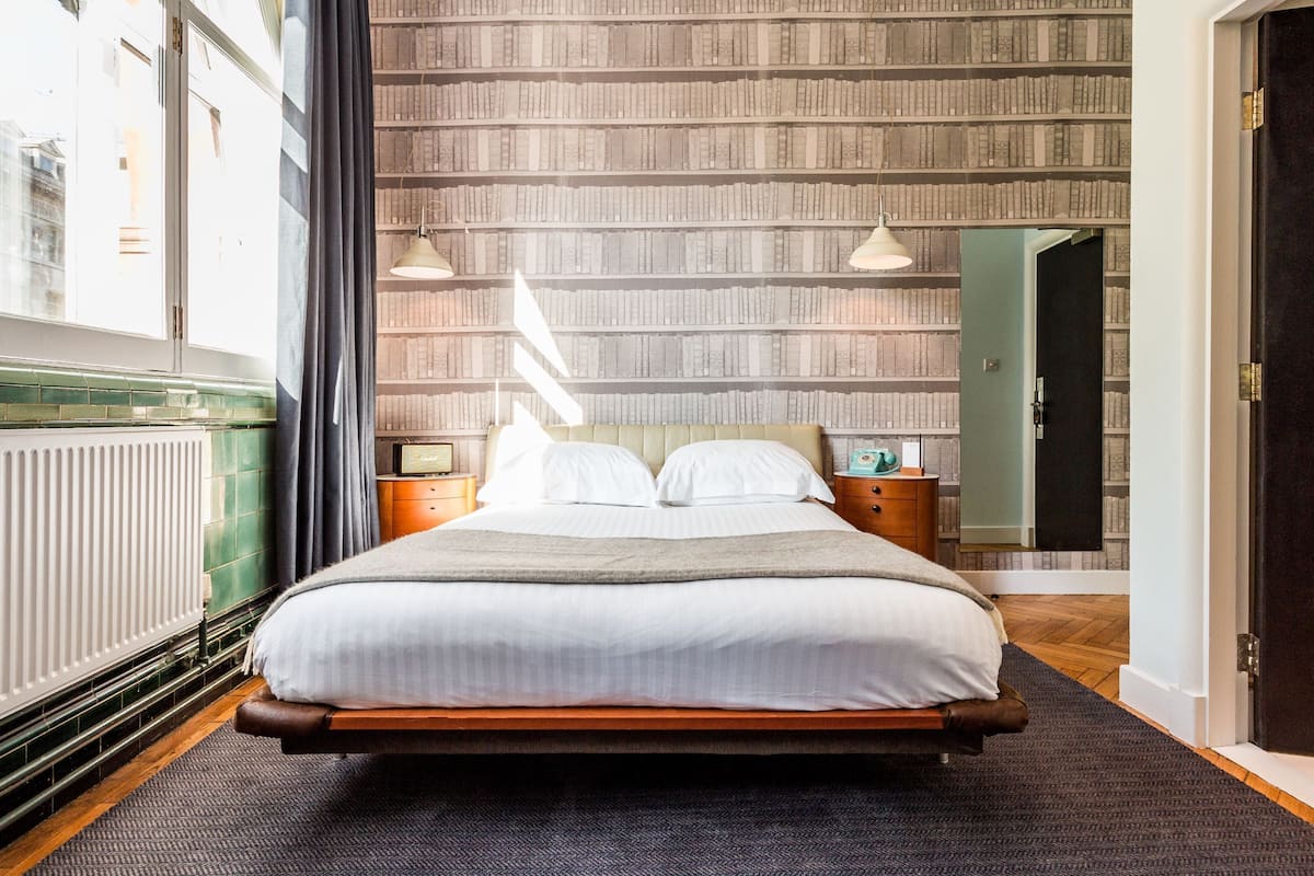 A neatly made bed with white linens, a wooden headboard, and a dark rug. There are two bedside tables with lamps, and a bookshelf filled with books is in the background.