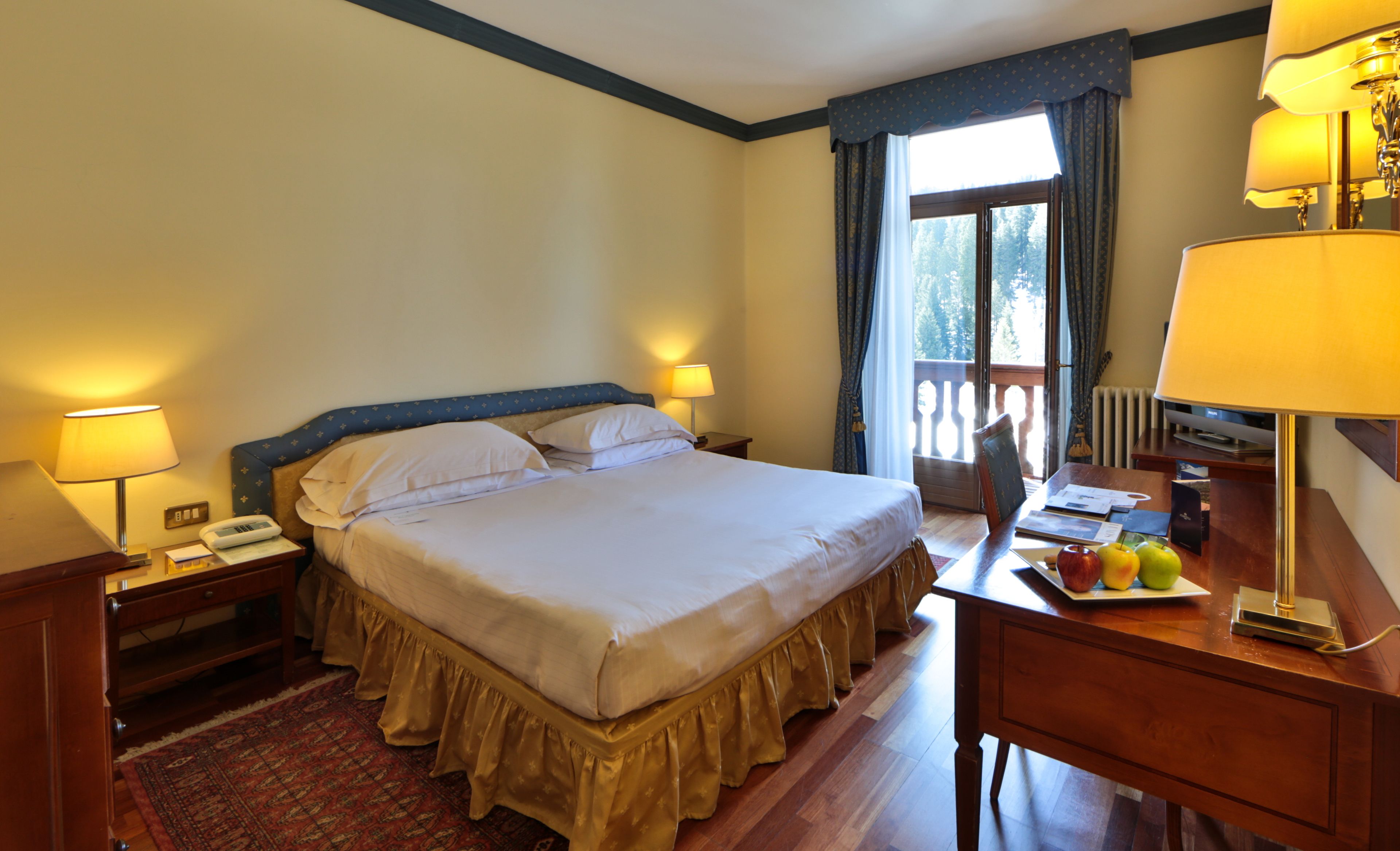 A hotel room with a large bed, two bedside lamps, a wooden desk with a lamp, a small table with fruit, and a view of the outdoors through a window.