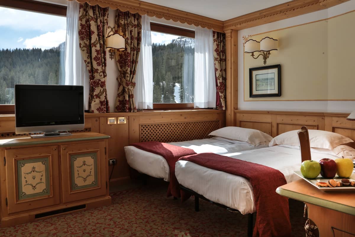 Habitación de hotel con dos camas, televisión, ventana con cortinas, cabecero de madera y una bandeja con fruta.