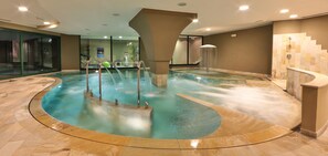 A large indoor swimming pool with a central water feature, surrounded by a curved walkway and a staircase leading to an upper level.