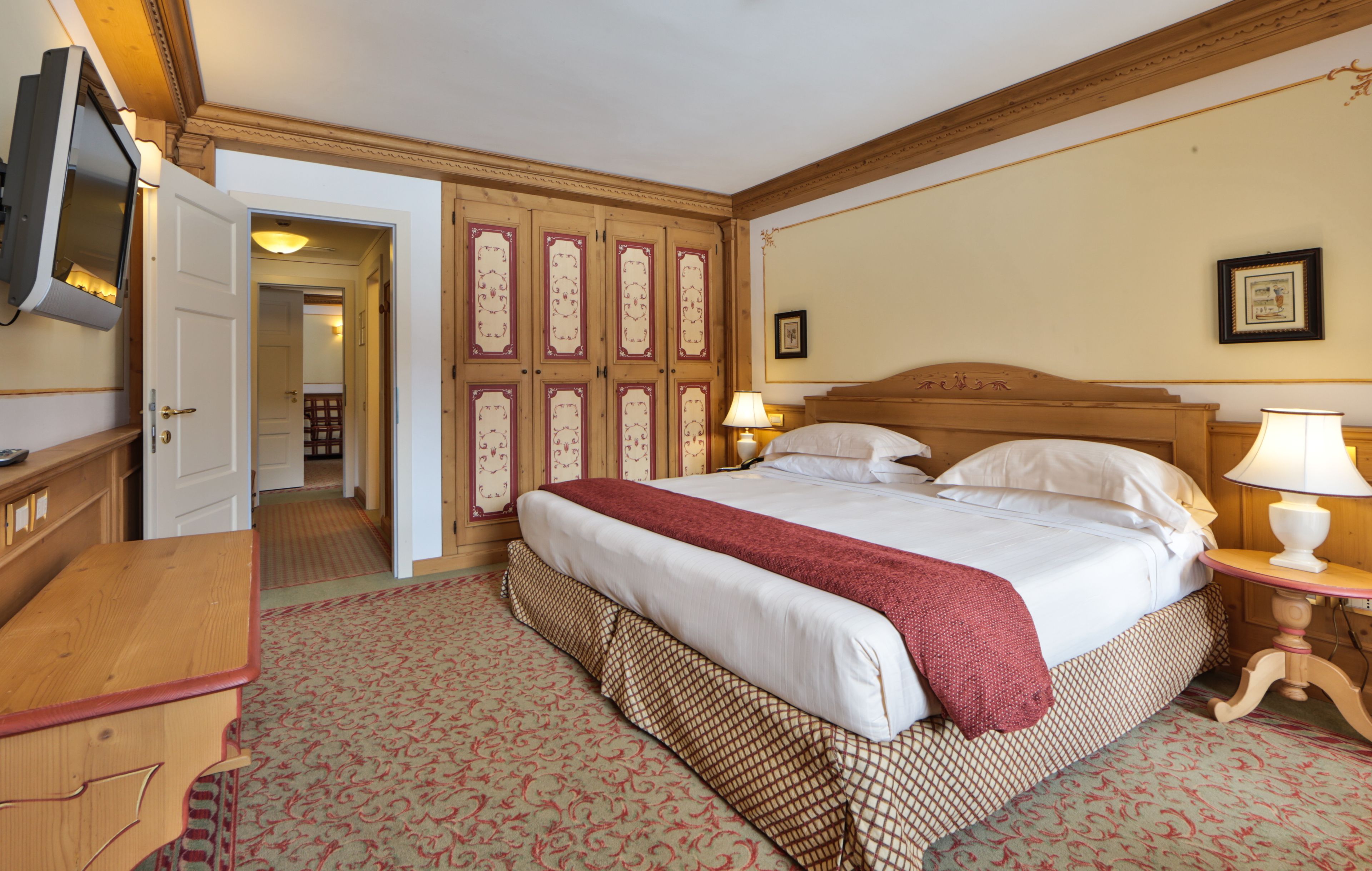 A hotel room with a large bed, a wooden headboard, a bedside table with a lamp, a television mounted on the wall, and a door leading to another room.