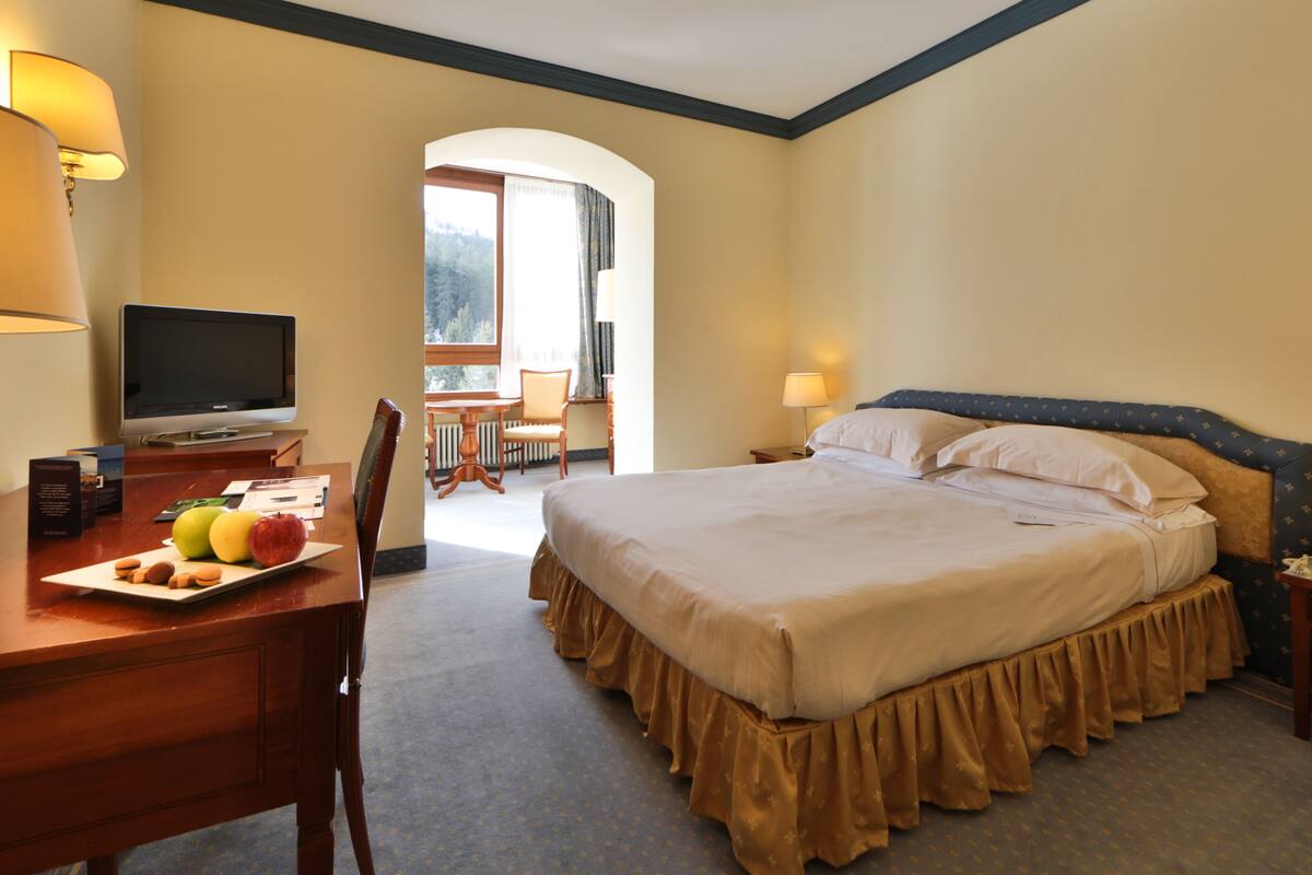 Habitación de hotel con una cama grande, un escritorio con una computadora, un televisor y una mesa de comedor con frutas y nueces.