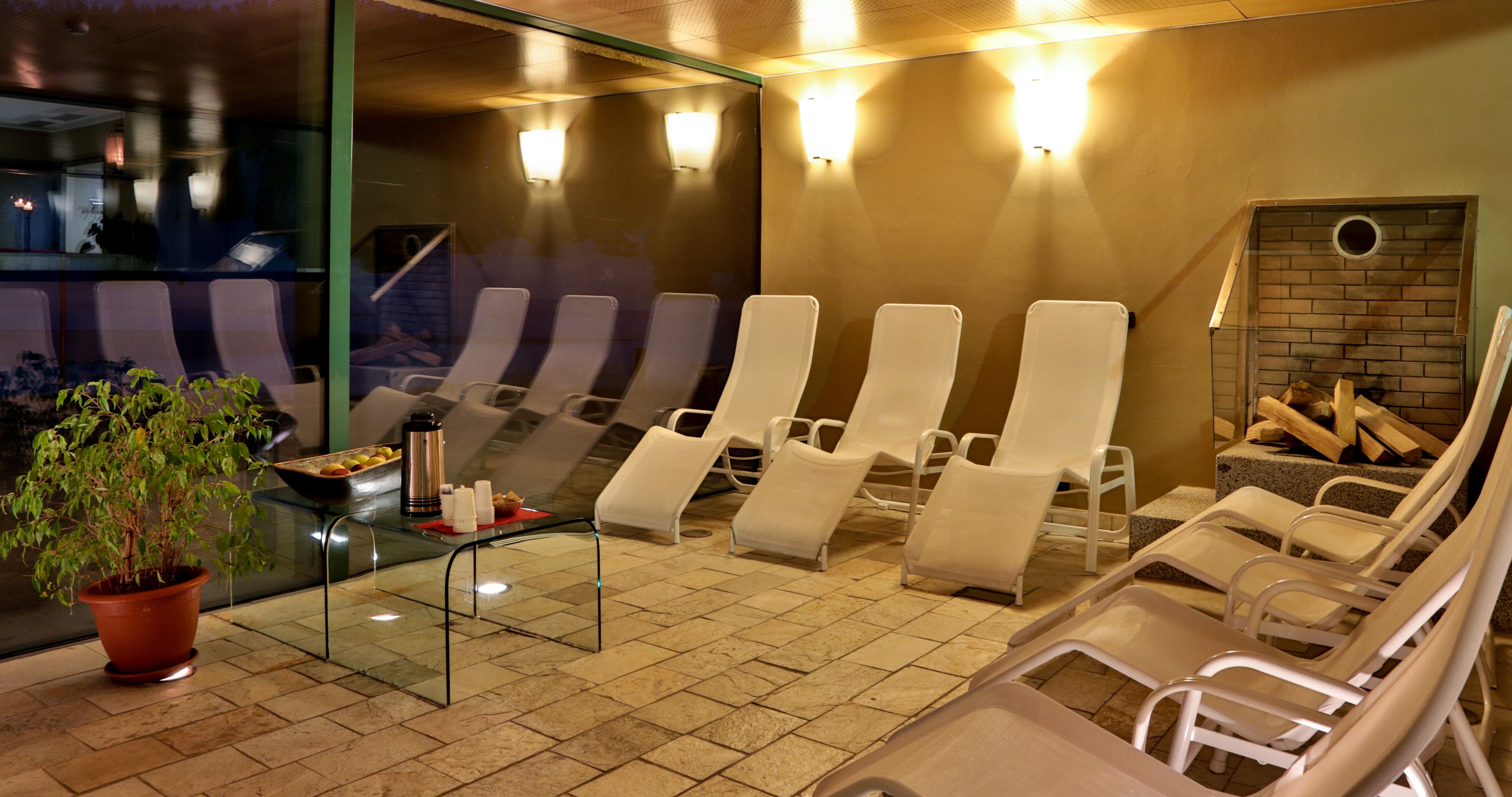 A spa area with reclining chairs, a glass table with a plant and a tray of fruits, and a brick wall with a circular window.