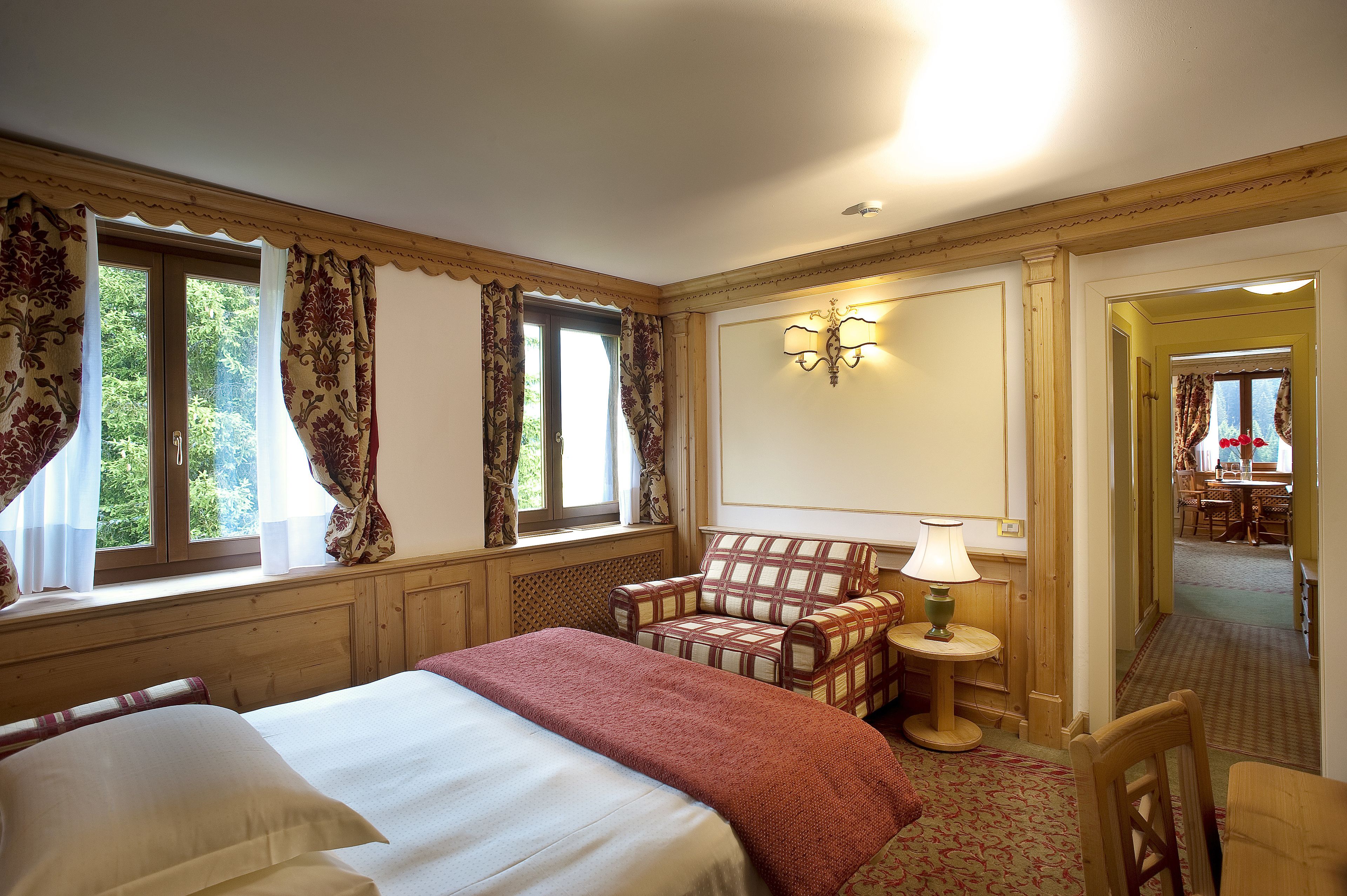 Ein Hotelzimmer mit einem großen Bett, einem Sessel, einem kleinen Tisch und einer Lampe. Ein Fenster mit Vorhängen und eine Tür zu einem weiteren Raum sind ebenfalls vorhanden.