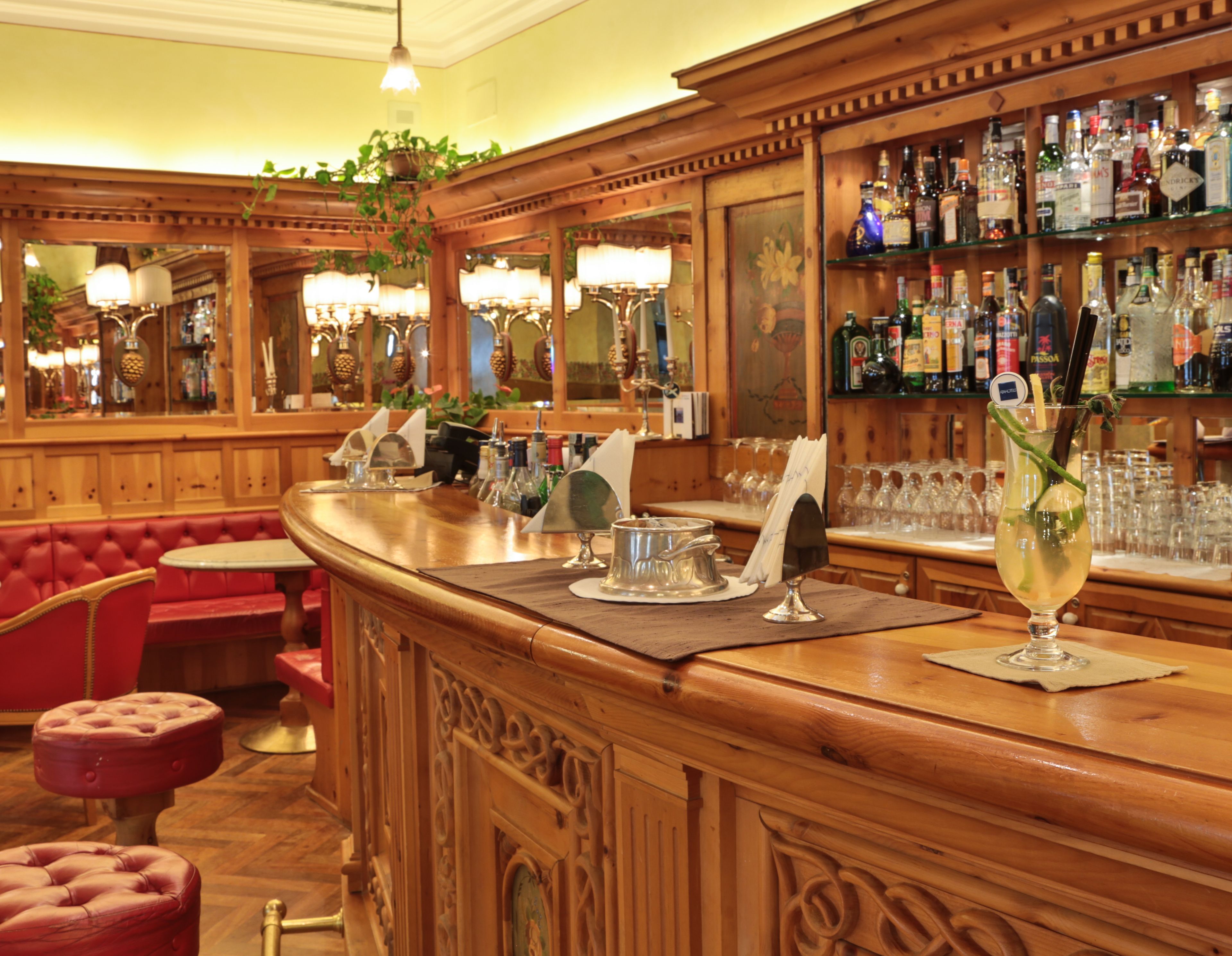 A bar area with a wooden counter, red cushioned seating, and a variety of bottles and glasses on shelves.