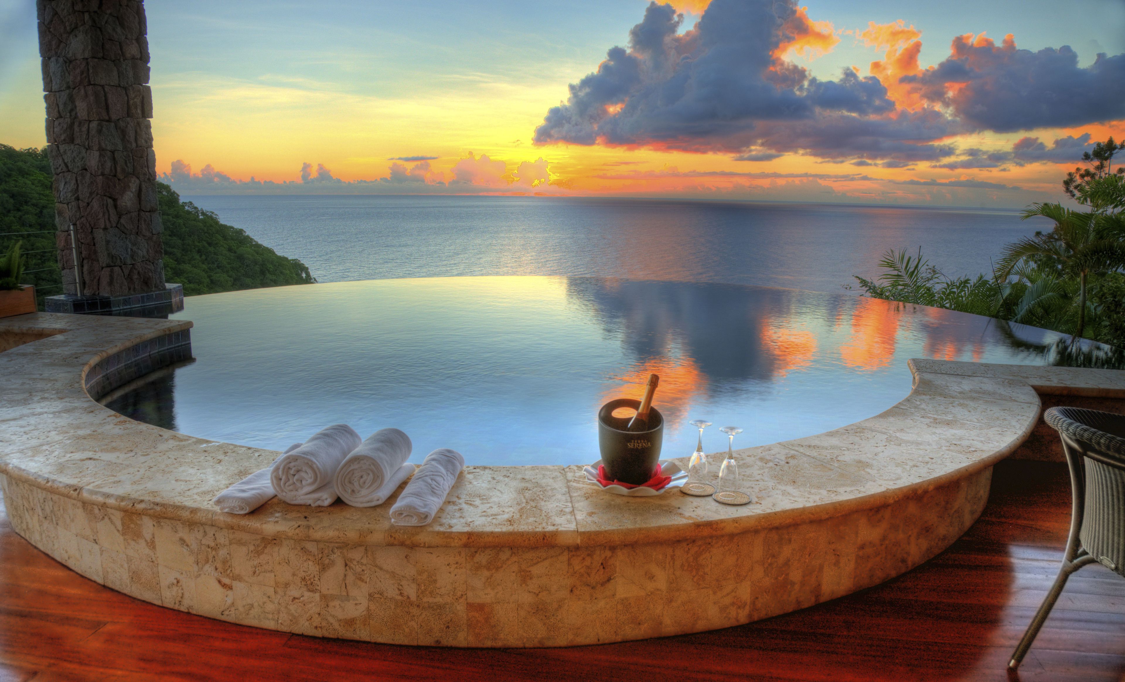 A circular infinity pool with a view of the sunset over the ocean, a stone pillar, and a small table with a bottle and glasses.