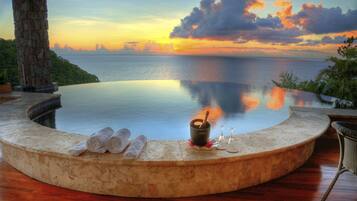 A circular infinity pool with a view of the sunset over the ocean, a stone pillar, and a small table with a bottle and glasses.