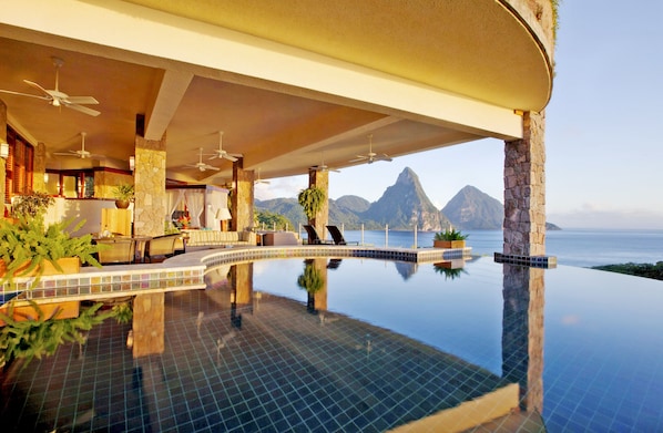 An infinity pool with a view of mountains and the ocean, a covered seating area with chairs and tables, and ceiling fans. - Jade Mountain Resort (Soufrière)