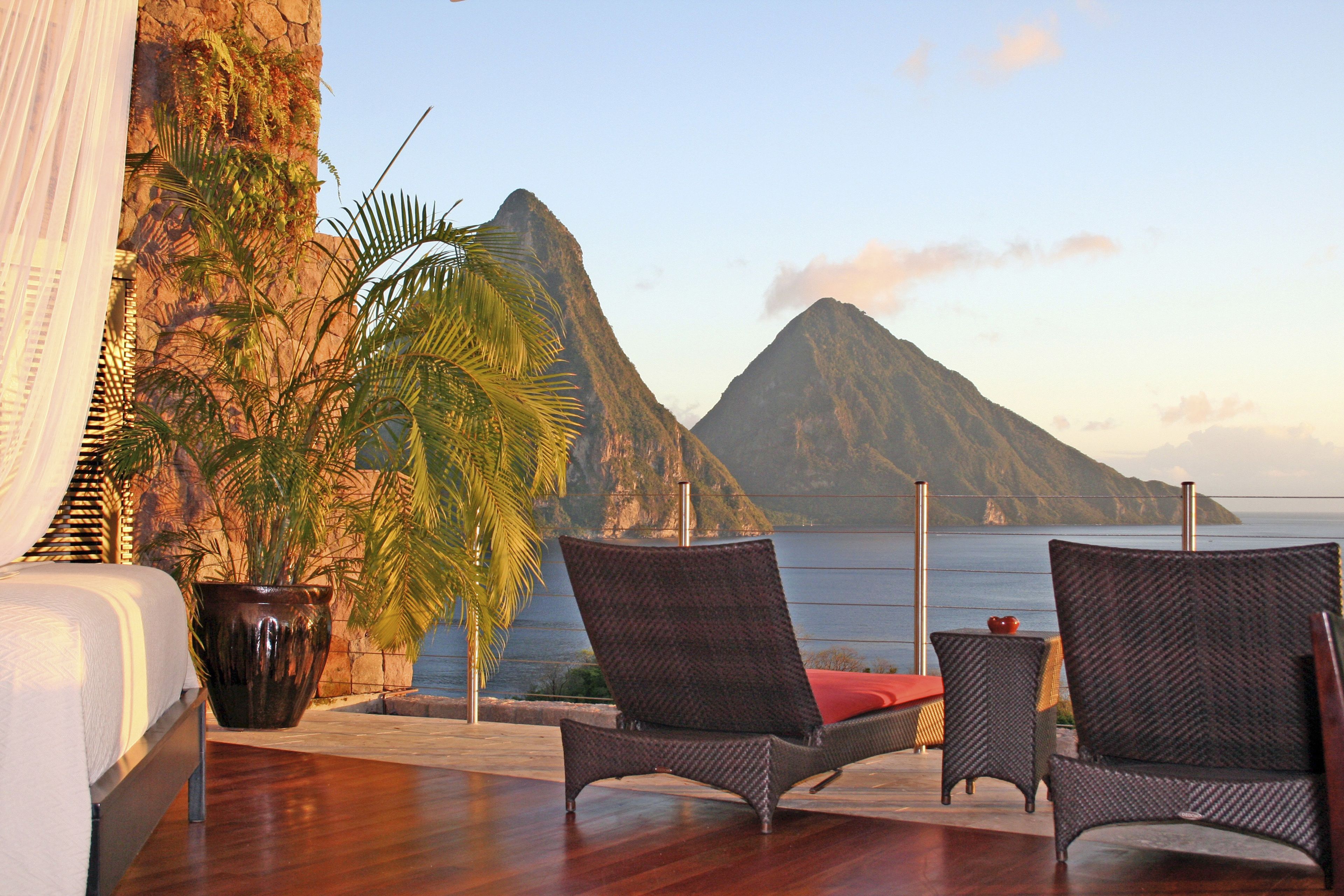 A balcony with wicker chairs, a potted plant, and a view of a mountain and the sea.
