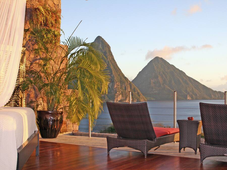 A balcony with wicker chairs, a potted plant, and a view of a mountain and the sea.