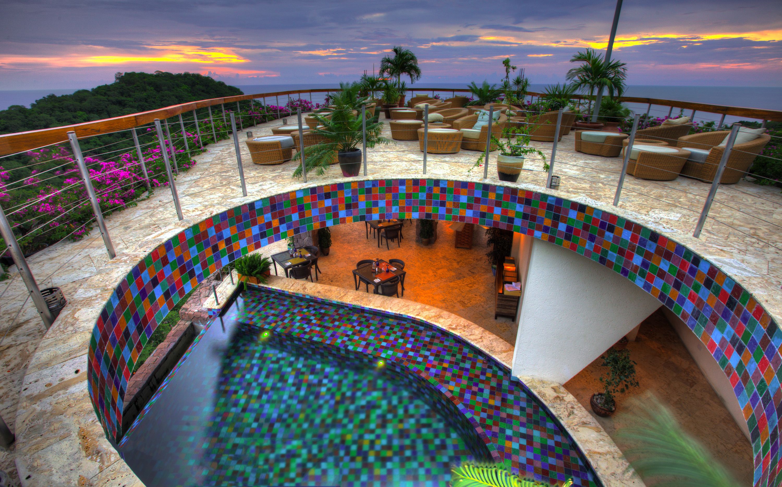 A rooftop pool area with a mosaic-tiled infinity edge, outdoor seating, and a view of the ocean at sunset.