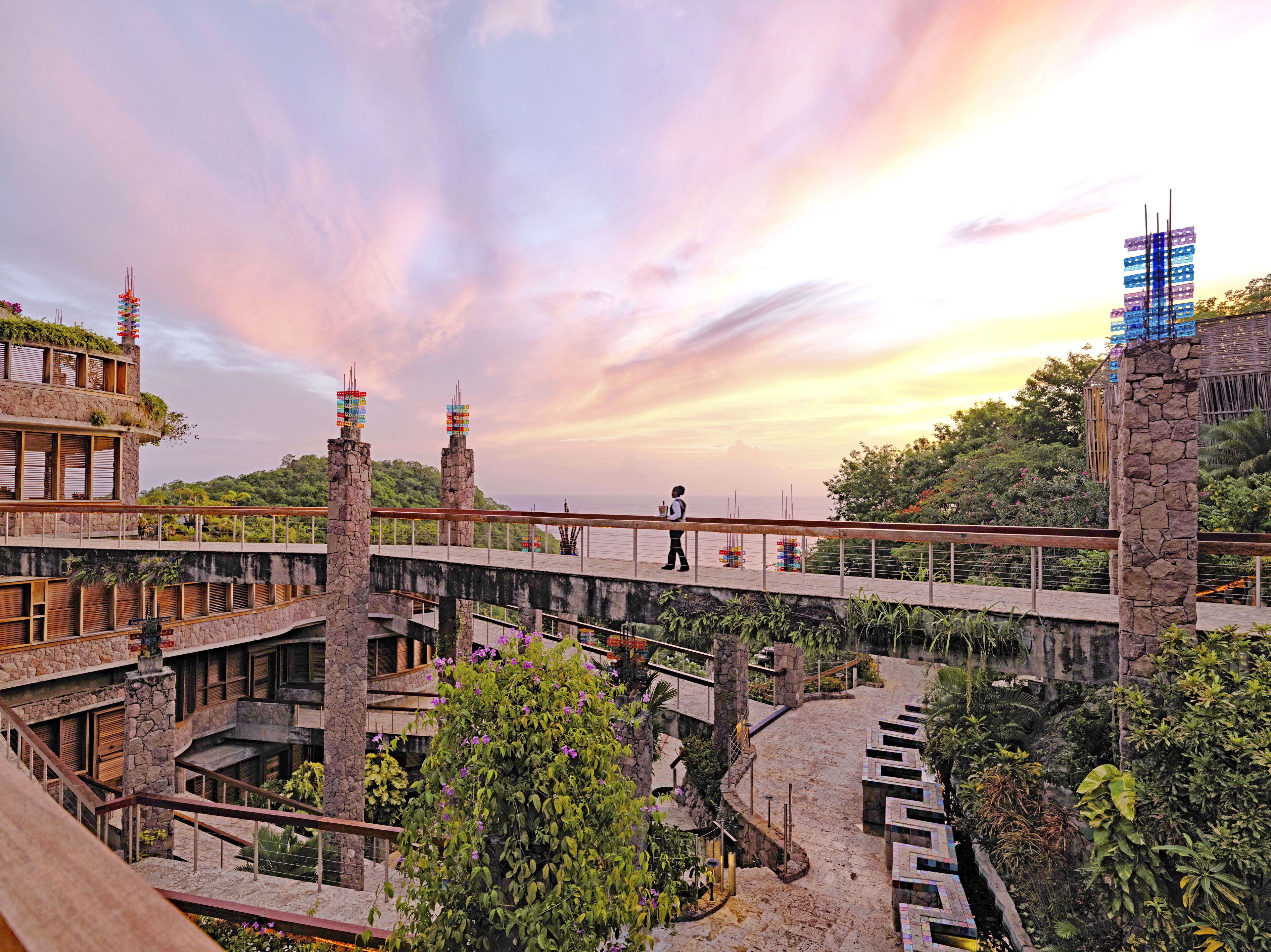 A multi-level hotel with a stone facade, multiple balconies, and a view of the sea at sunset.